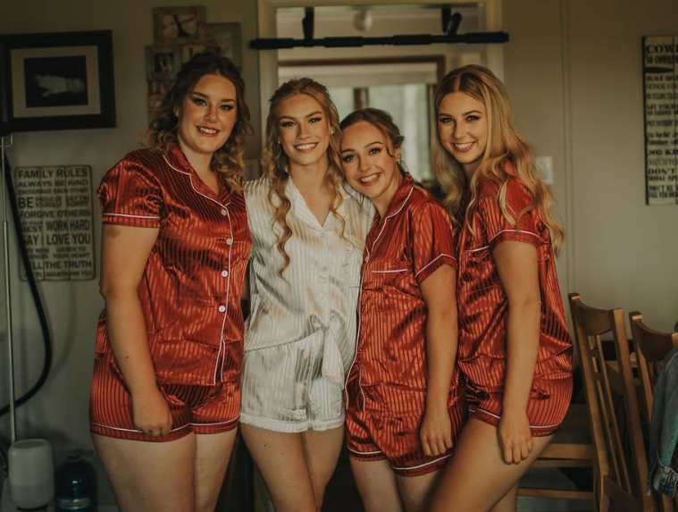 Three bridesmaids and the bride in their pajamas smiling and posing together in a cozy room with a mirror, framed pictures, and dining chairs in the background.