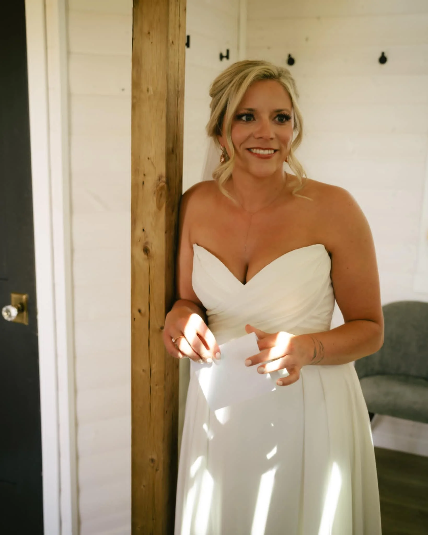 A woman in a white strapless dress smiling while holding a piece of paper, standing beside a rustic wooden post, with a white wall with hooks in the background.