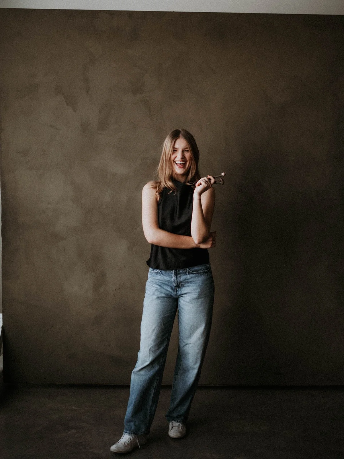 A young woman with light brown hair smiling, holding a small bouquet of flowers, wearing a black sleeveless top and light blue jeans, standing against a plain, dark wall.