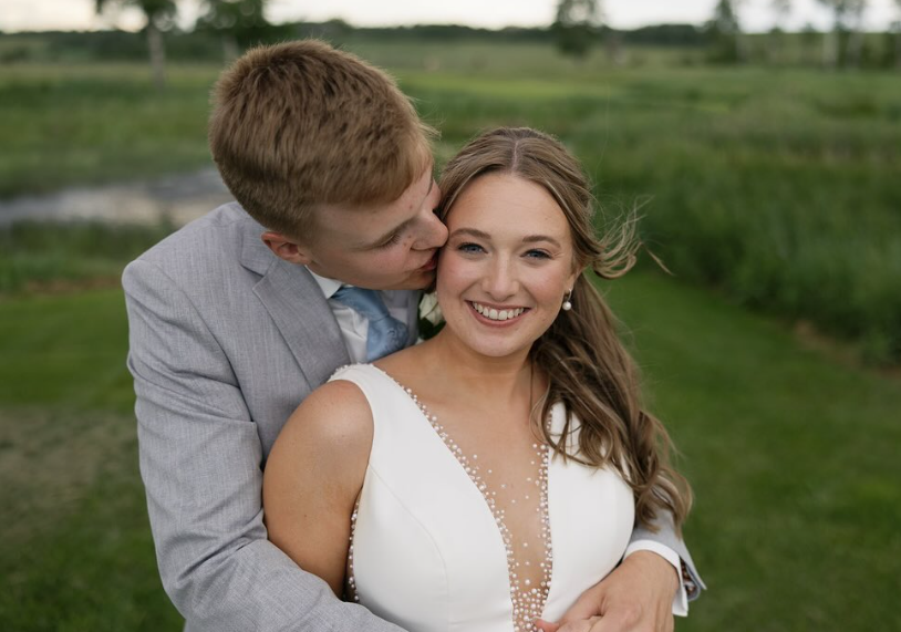 A man in a gray suit kisses a smiling woman in a white dress outside in a grassy area.