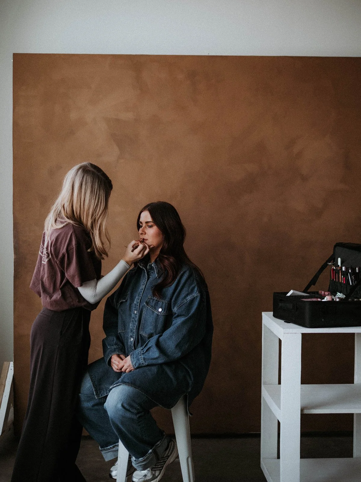 A woman getting her makeup done by a makeup artist at a photoshoot studio. The makeup artist is applying makeup to the woman who is sitting on a high chair against a brown backdrop. A makeup kit with brushes and cosmetics is on a white table to the right.