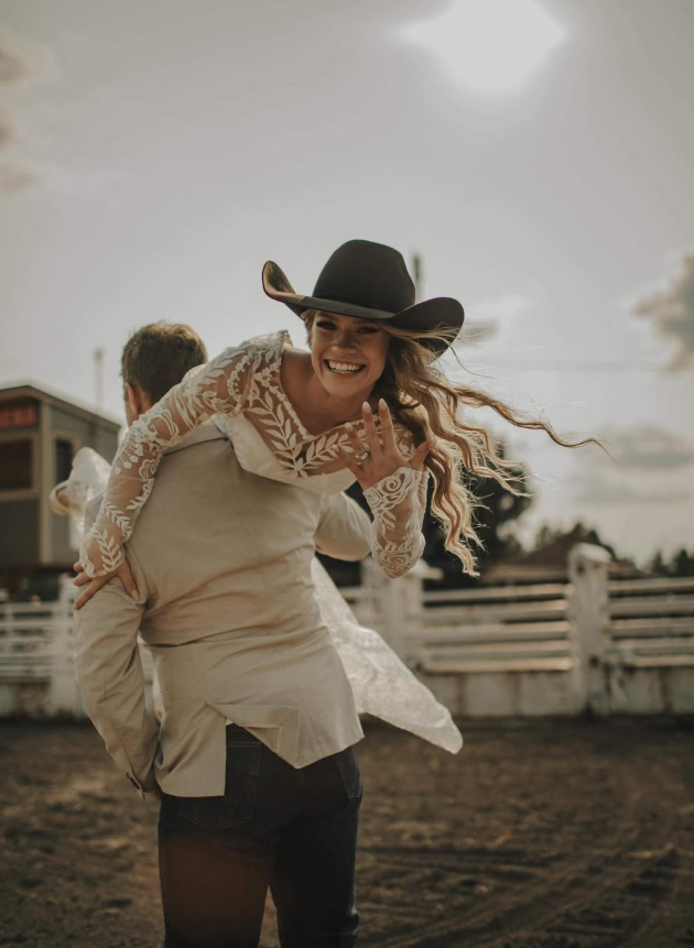 Woman in a white dress and black cowboy hat happily piggybacking on a man at a farm or ranch.