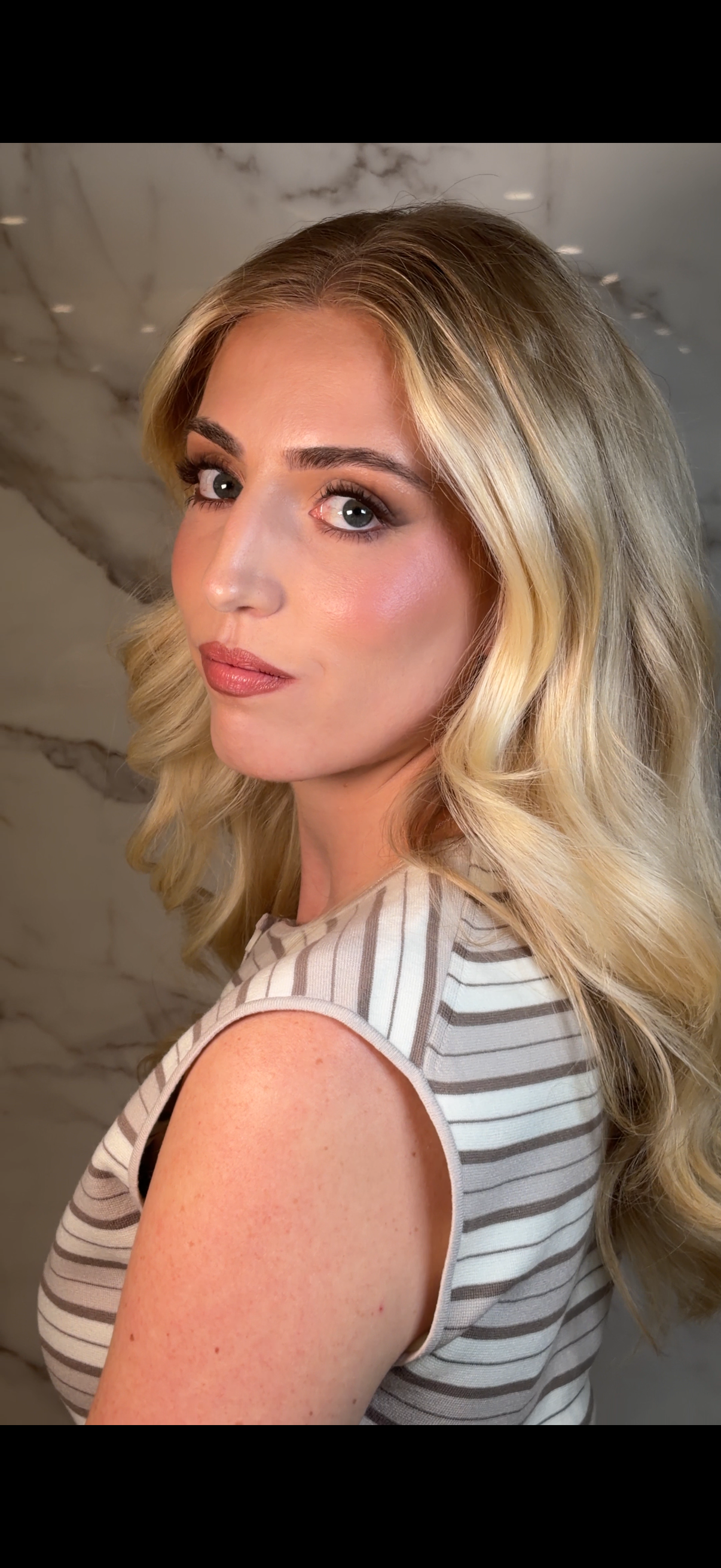 Close-up of a woman with blonde wavy hair, smoky eye makeup, and pink lipstick, wearing a striped sleeveless top, standing against a marble background.