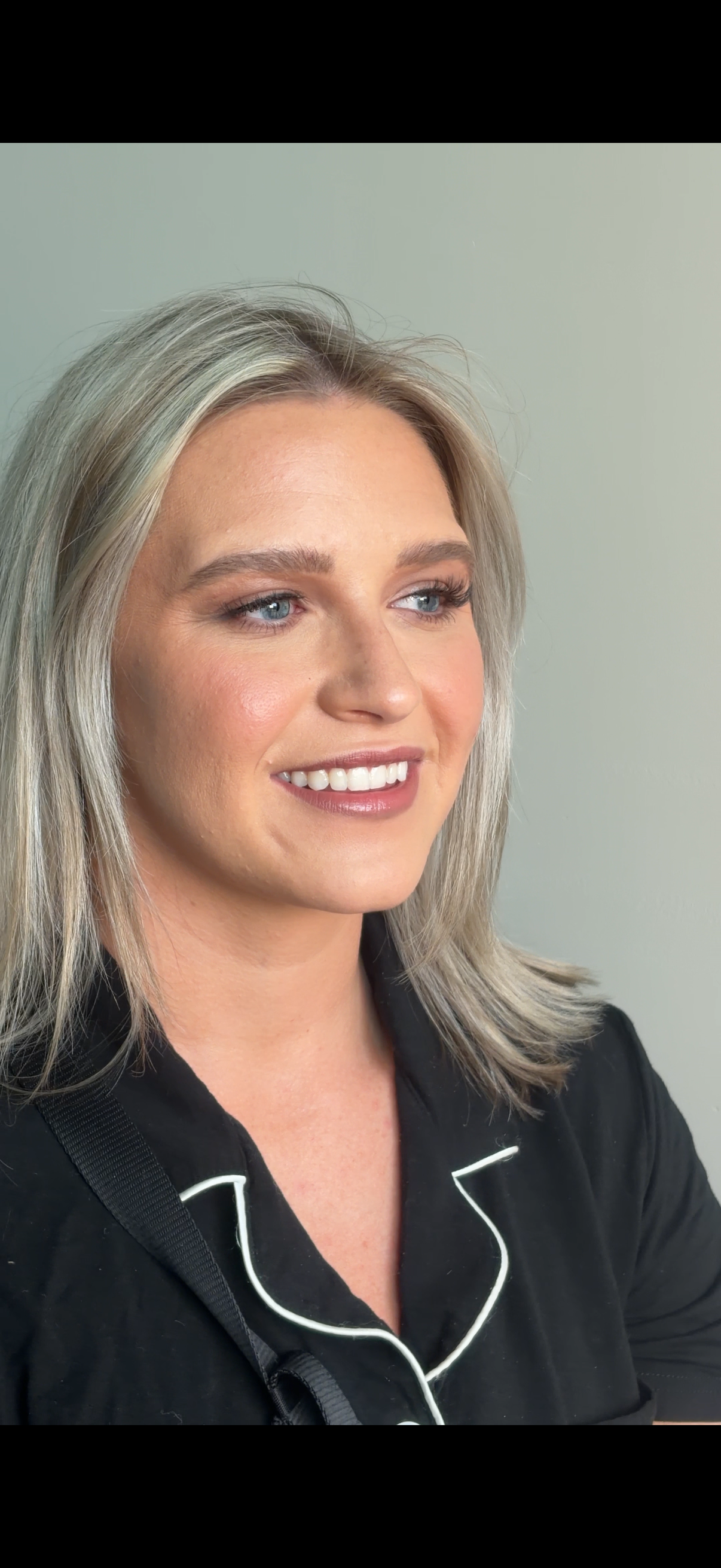 Close-up photo of woman with blonde hair, blue eyes, smiling, wearing black pajamas with white piping, against light-colored background.