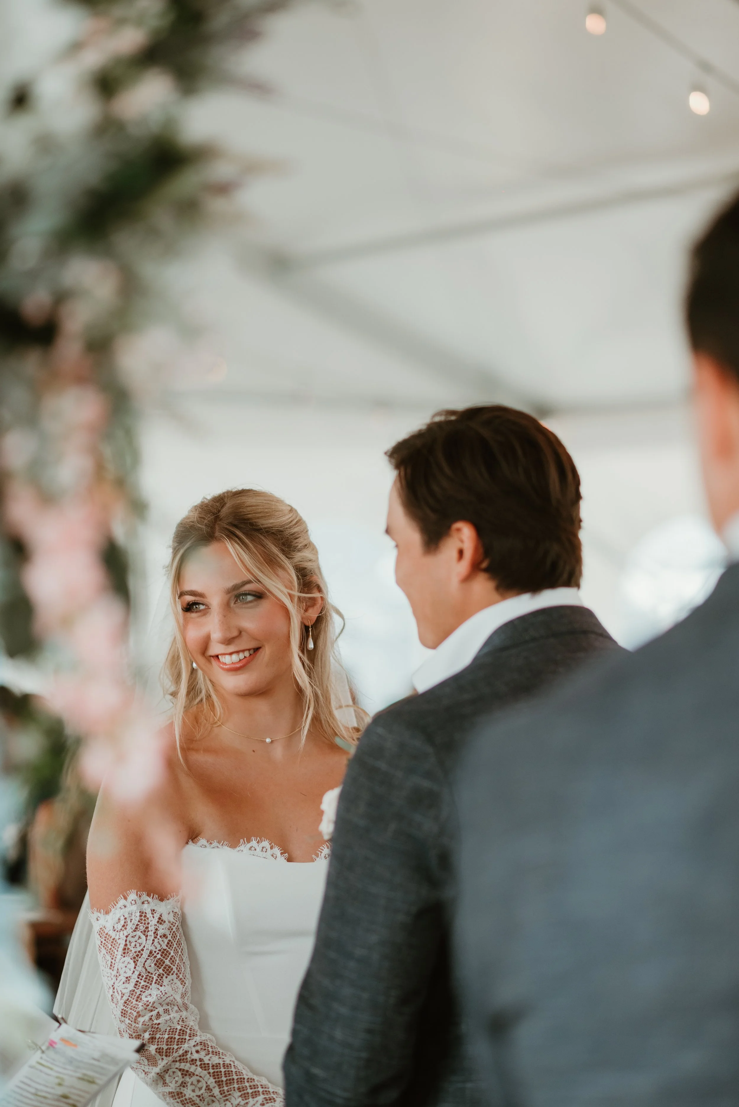 A bride with blonde hair and blue eyes, wearing a white wedding dress with lace sleeves, looks at her groom during their wedding ceremony. The groom has dark hair and is dressed in a dark suit. They are under a decorated canopy with greenery and pink