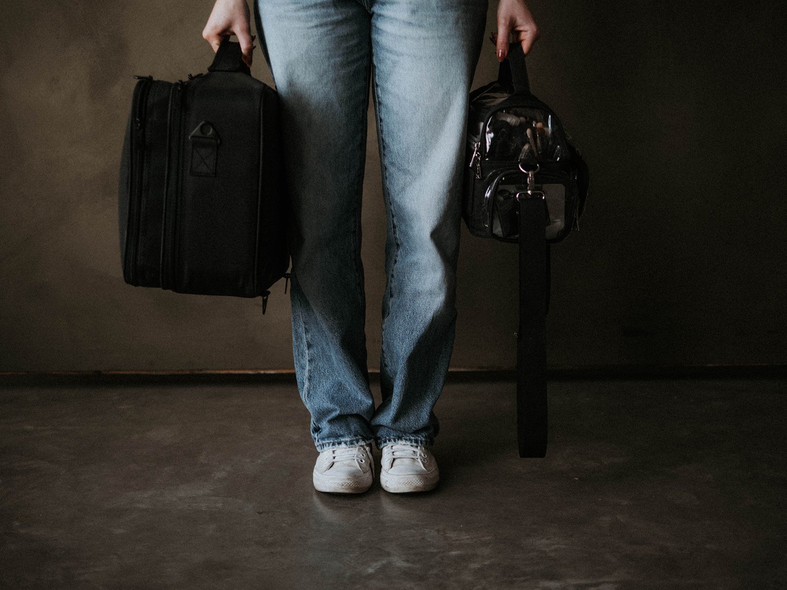 Person standing on a dark floor, jeans and white sneakers, holding a black rolling suitcase in the left hand and a small black handbag with makeup brushes inside in the right hand.