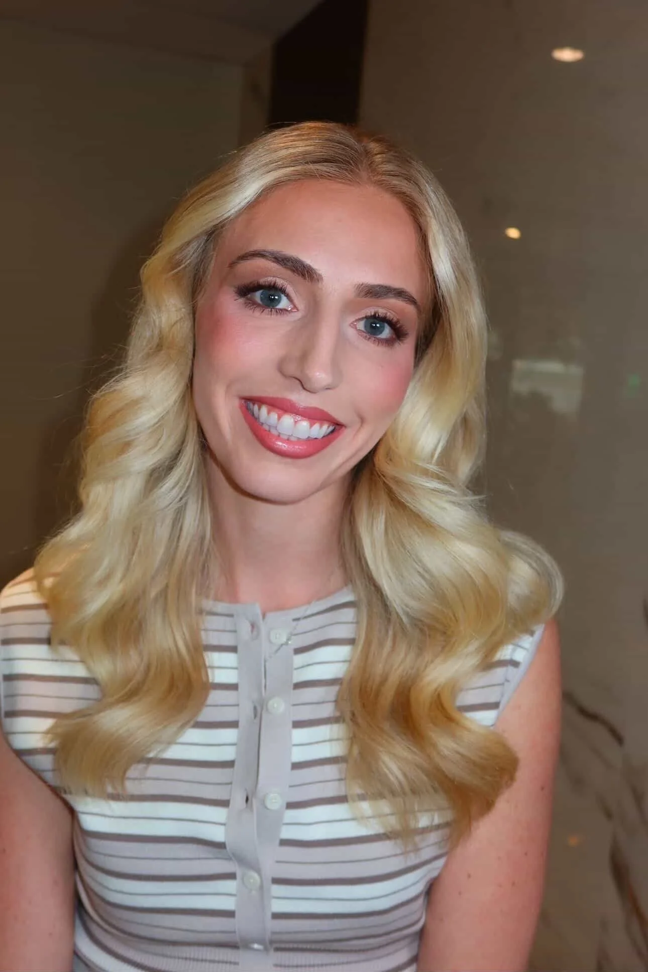 A smiling woman with long, wavy blonde hair, blue eyes, and wearing a sleeveless striped shirt.