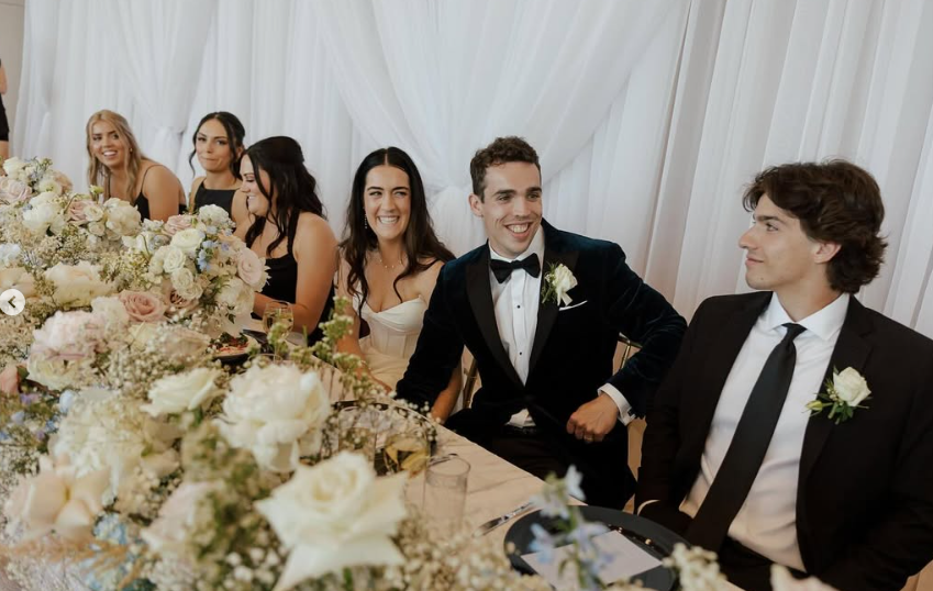 People in formal attire at a wedding reception, sitting at a decorated table with flowers, white curtains in the background