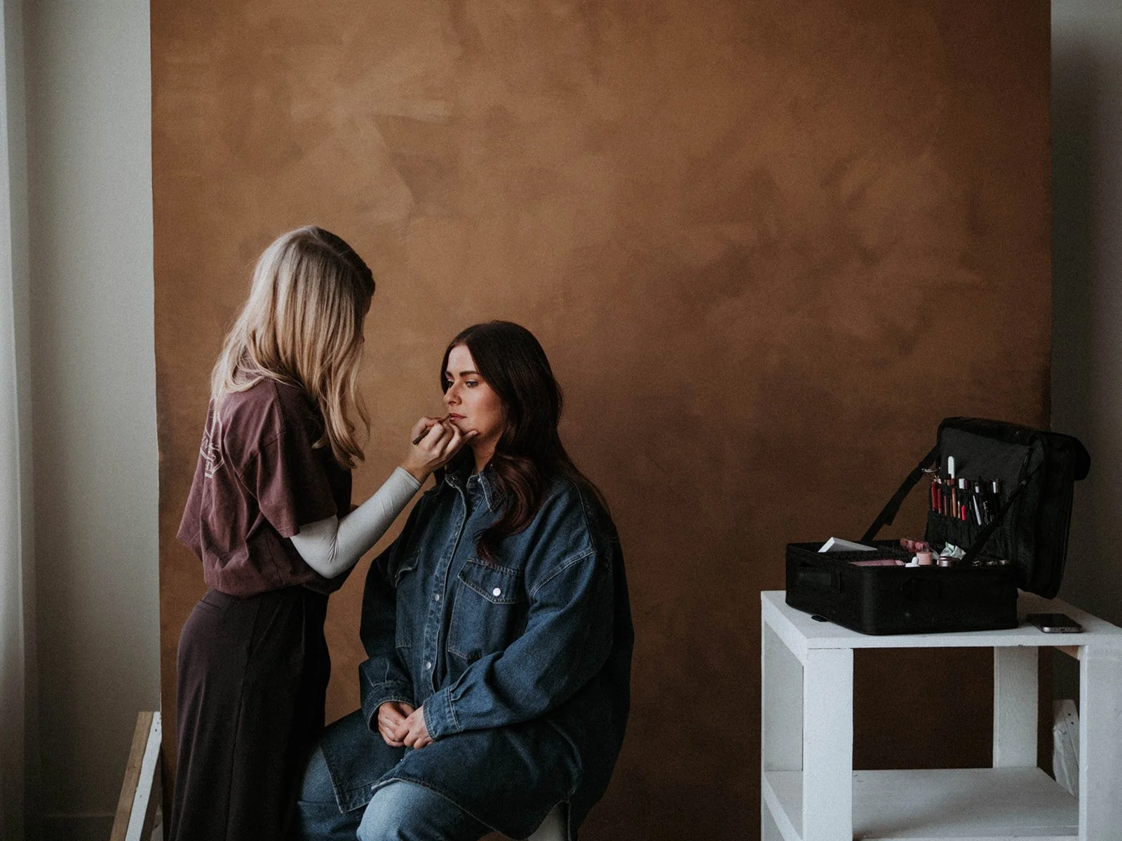 A woman applying makeup to another woman seated in front of a brown wall, with a makeup kit open on a white table nearby.