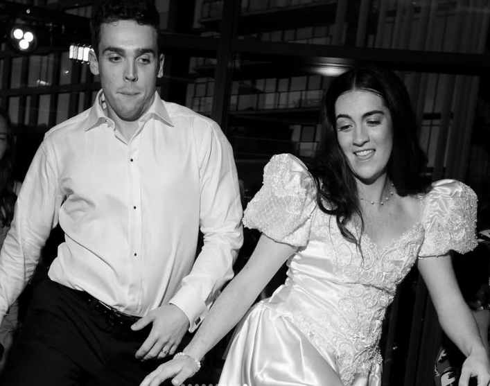 Black and white photo of a young man and woman dancing together at a formal event. The man is wearing a white shirt and dark pants, and the woman is wearing a white dress with puffed sleeves.