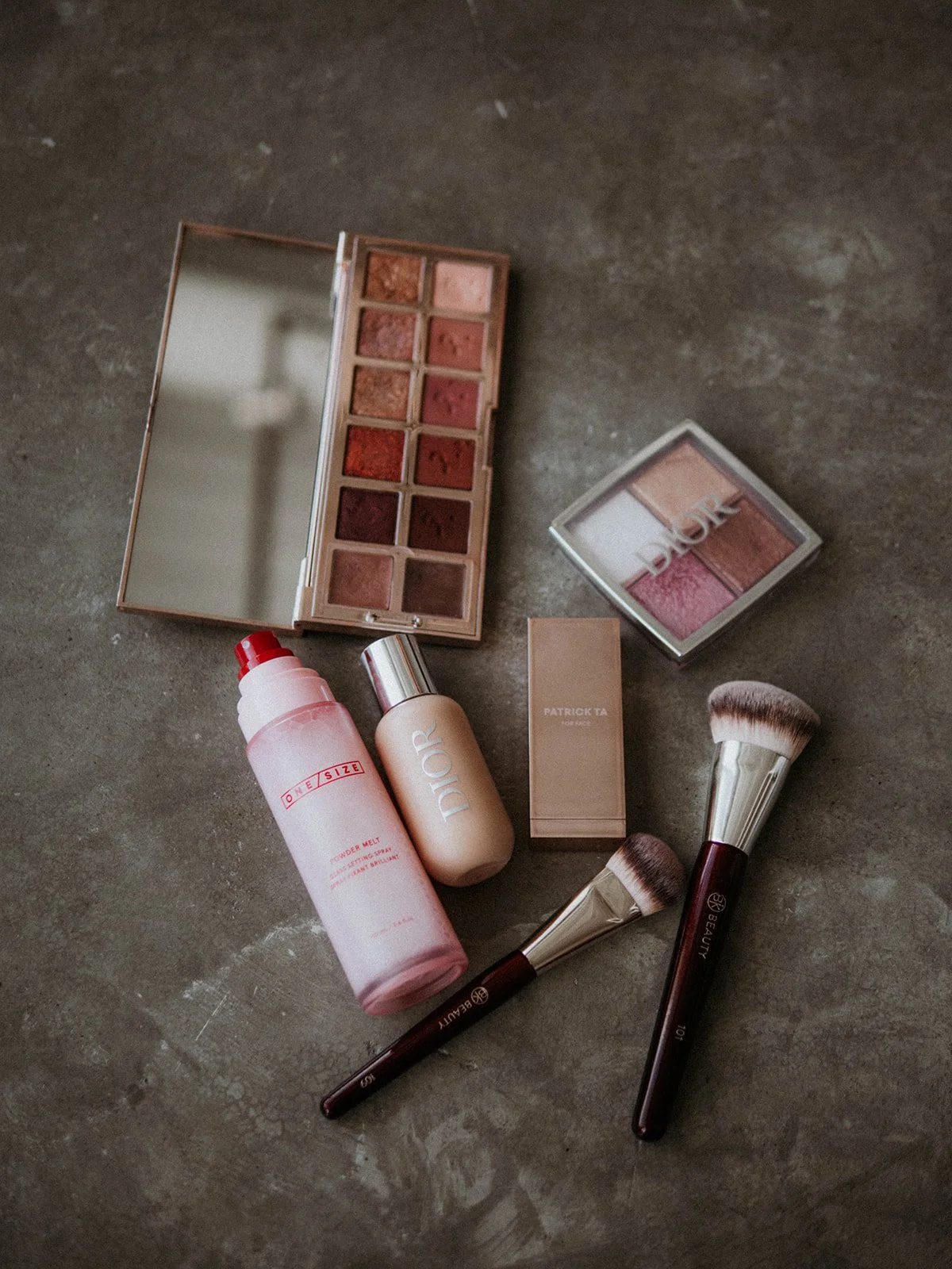 Makeup products including two large brushes, a facial mist, a foundation bottle, a beige lipstick, and two eyeshadow palettes with neutral and pink shades, all arranged on a gray surface.