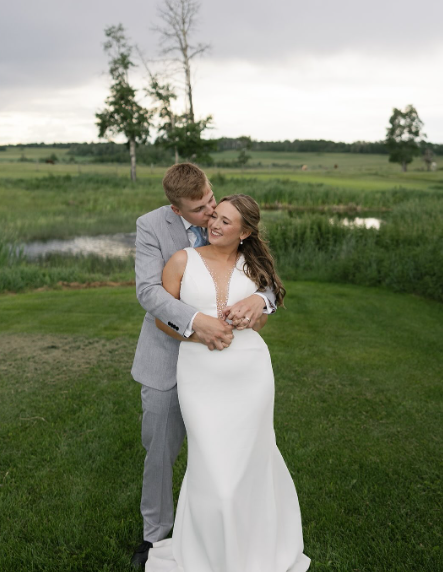 A newlywed couple embracing outdoors on a grassy field with a pond and trees in the background.