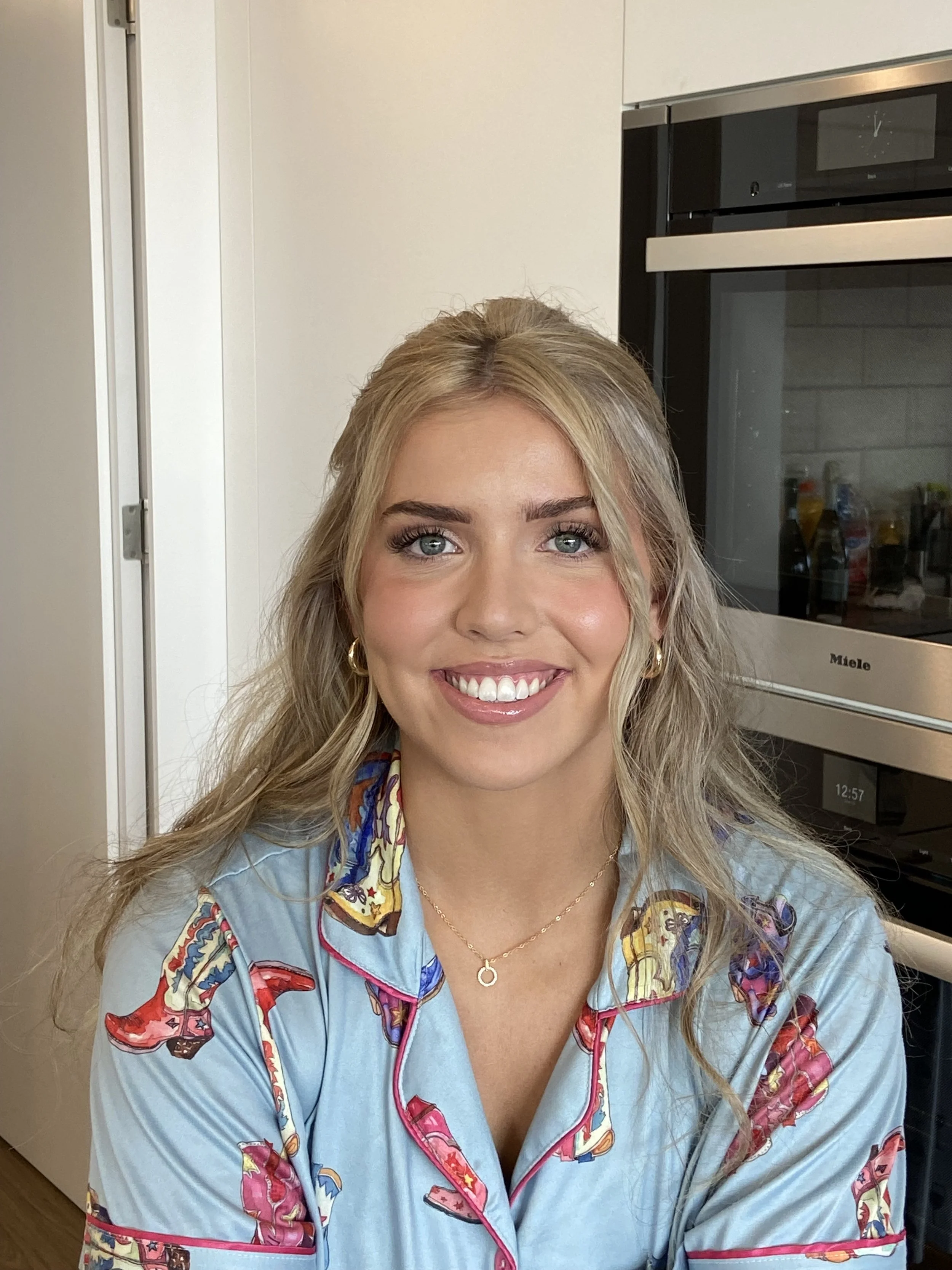 A smiling woman with blonde wavy hair and blue eyes, wearing a colorful patterned robe, gold hoop earrings, and a gold necklace, in a kitchen setting.