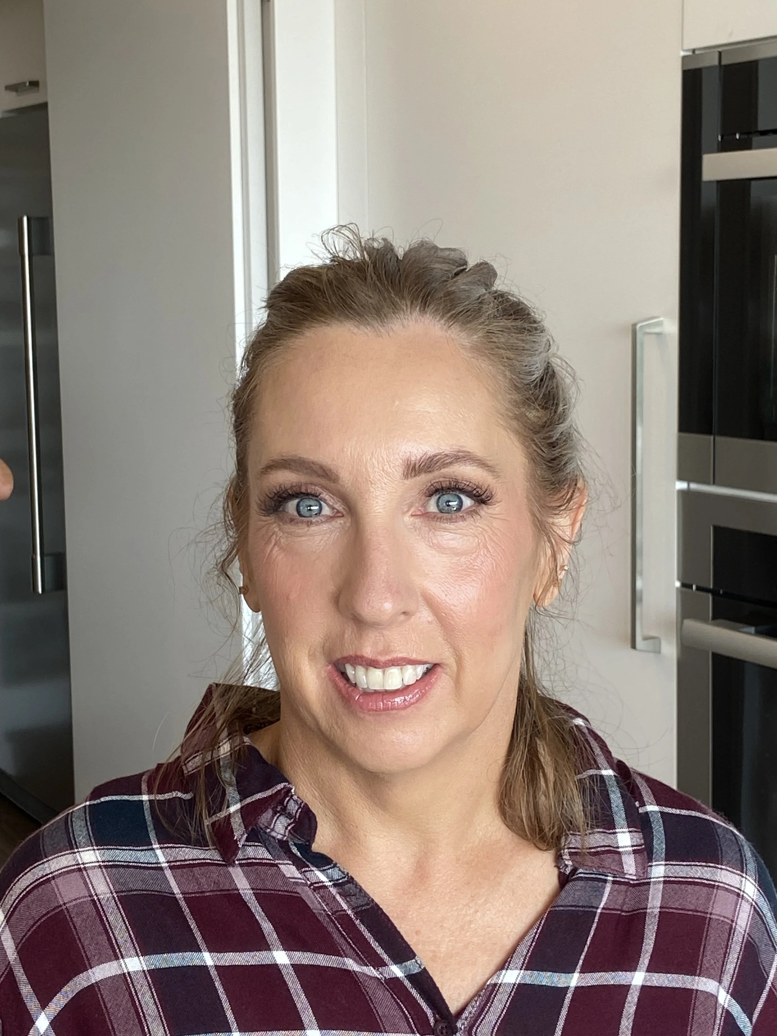 A woman with blue eyes and light brown hair styled in a half-up ponytail, smiling at the camera, wearing a purple plaid shirt, in a modern kitchen setting.