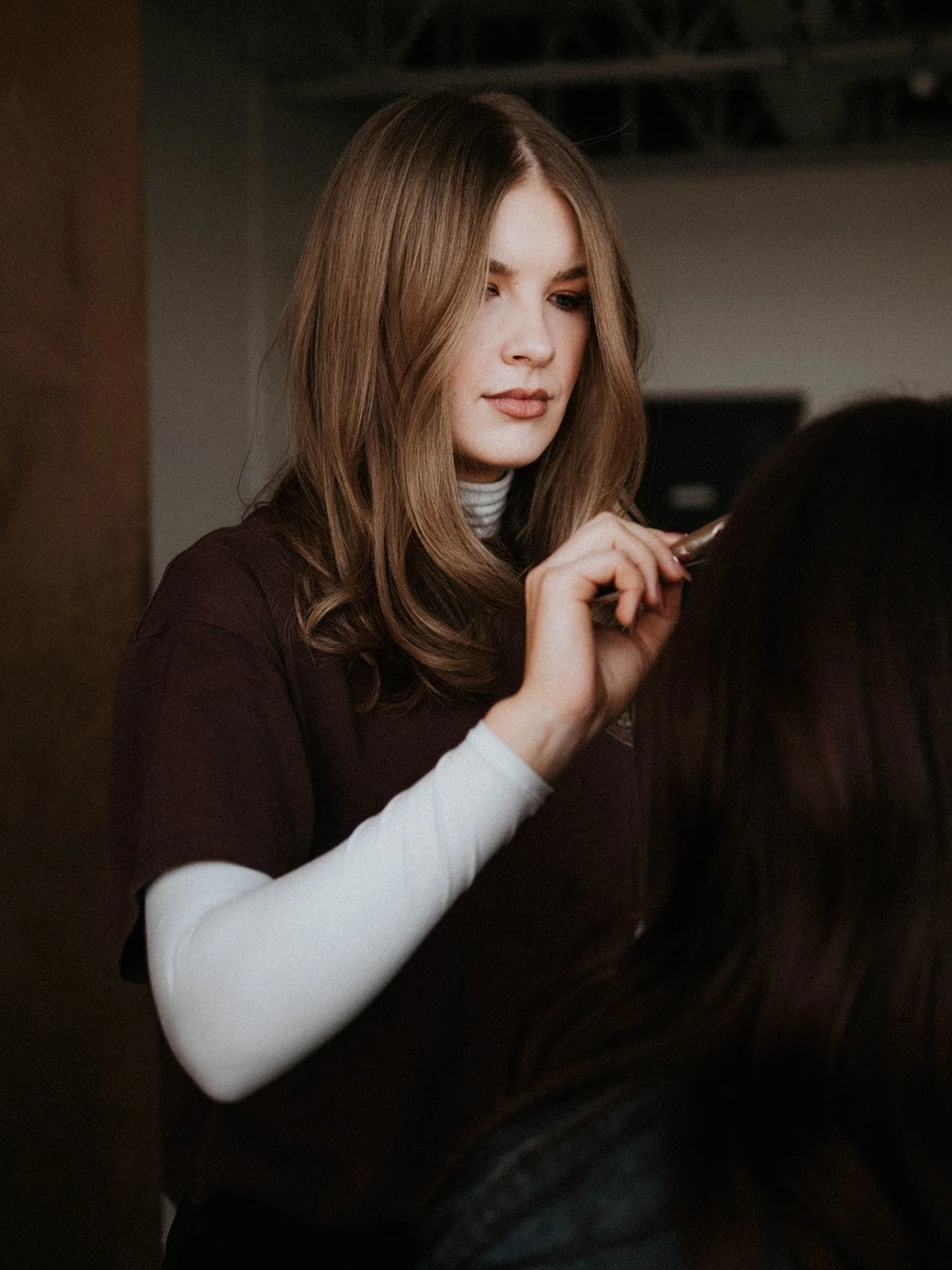 A woman with long, wavy brown hair is holding a hairbrush and is styling another person's long, dark hair.
