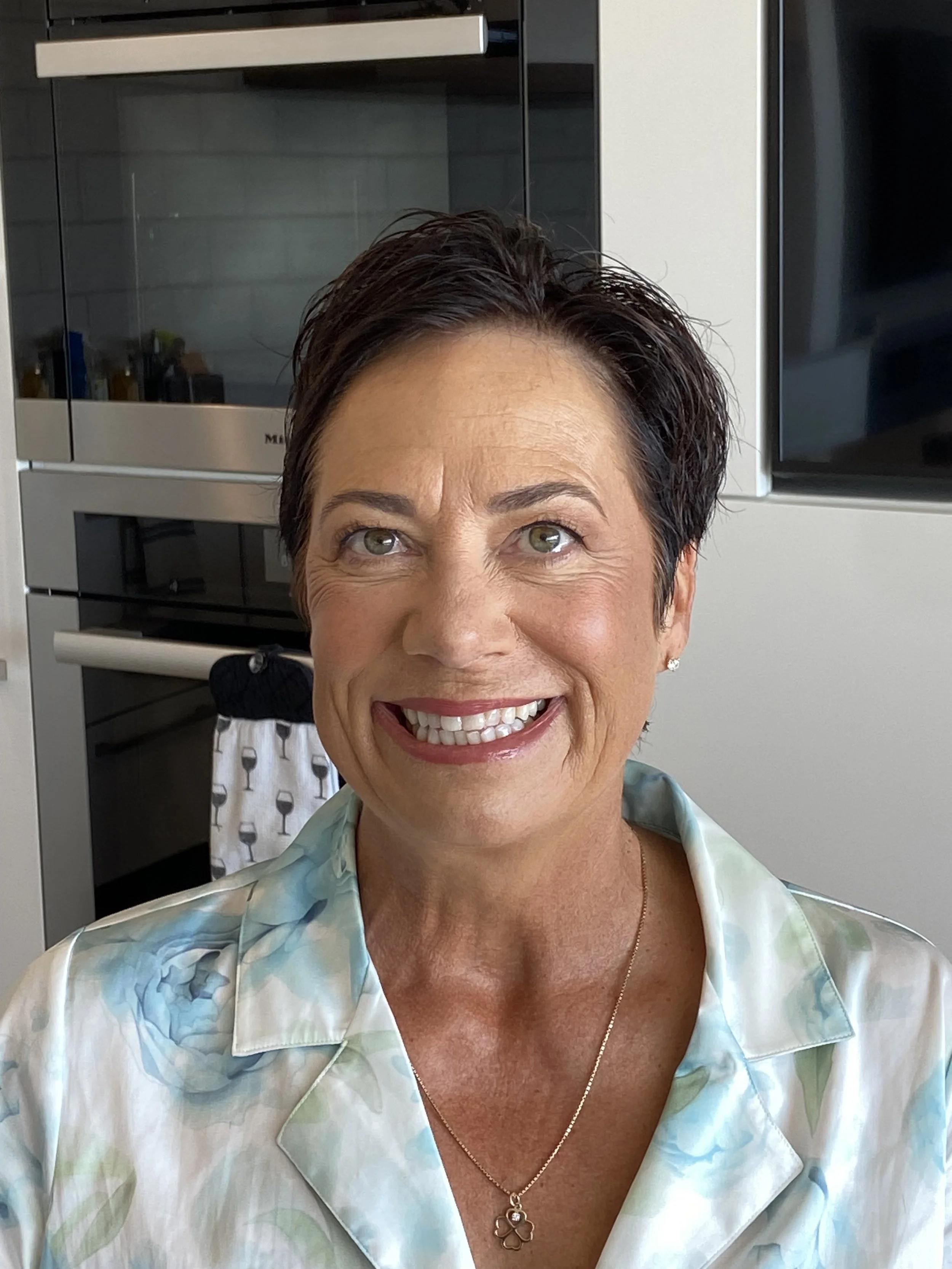 Smiling middle-aged woman with short dark hair, wearing a pastel-colored blouse and a clover-shaped pendant necklace, sitting in the kitchen with oven and microwave in the background.
