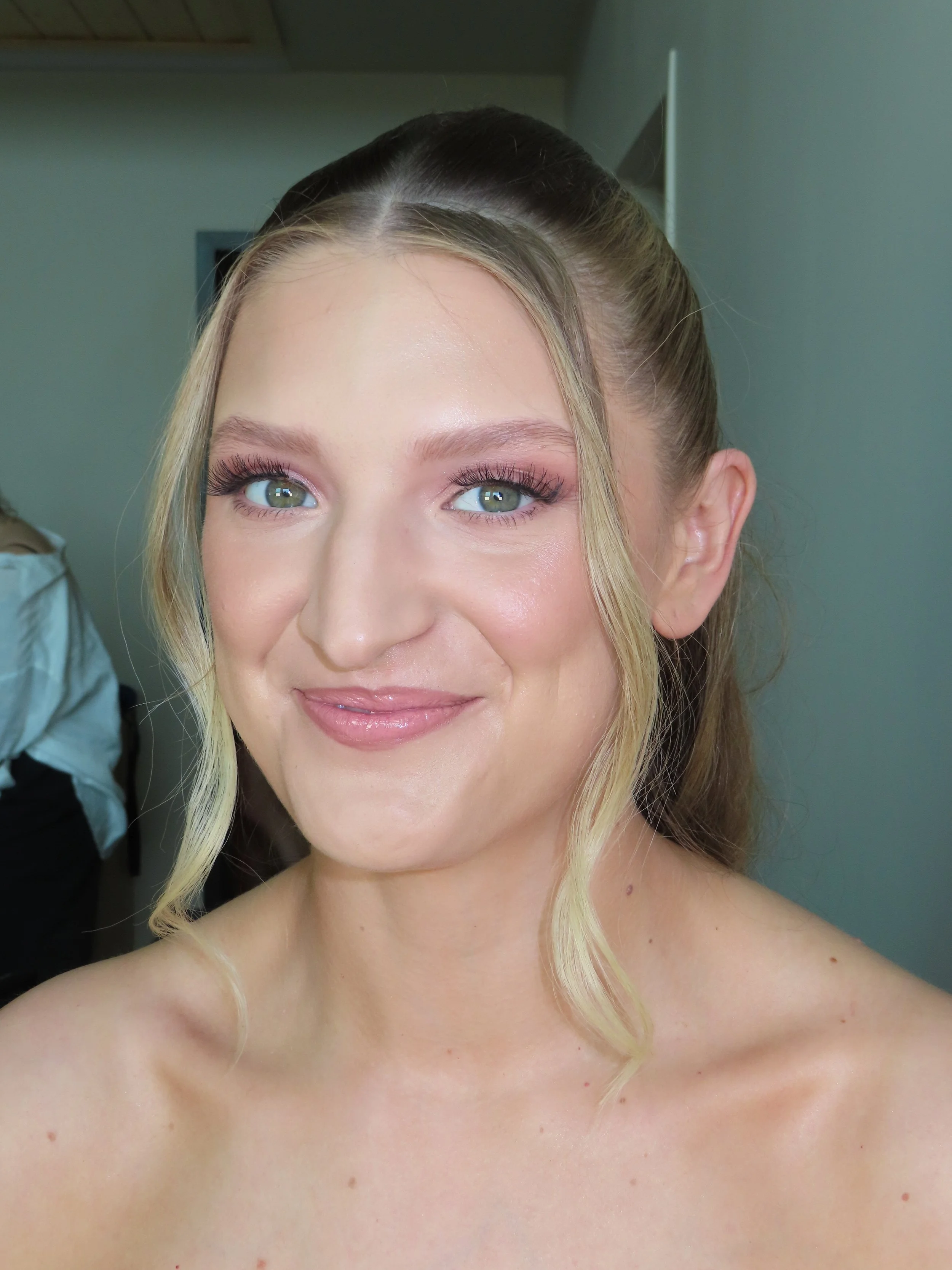 Close-up of a smiling woman with blond hair and light eyes, wearing minimal makeup and no clothing visible.