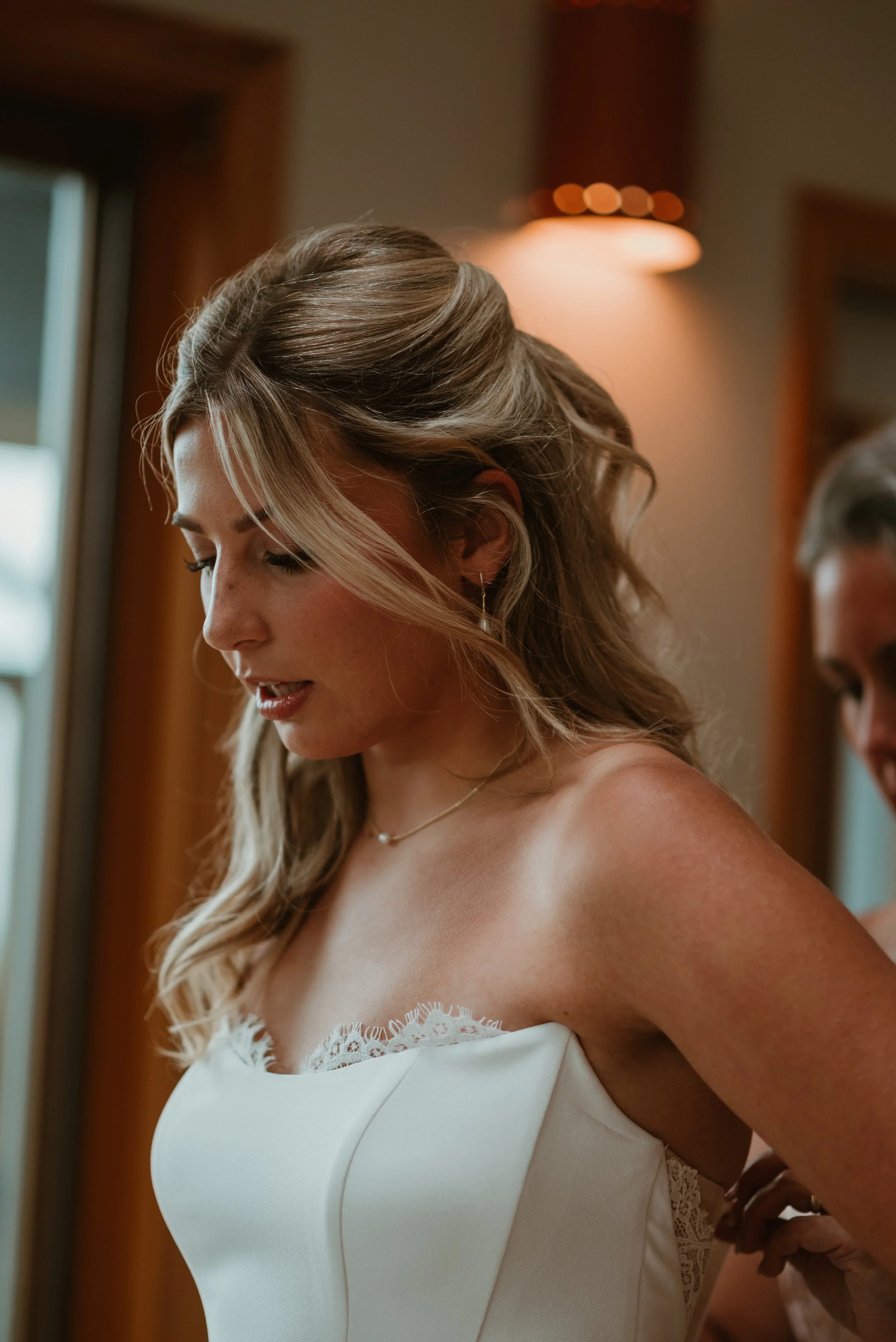 A woman with styled brown hair in a bridal gown with lace details, standing indoors near a window and a wall light, looking downward.