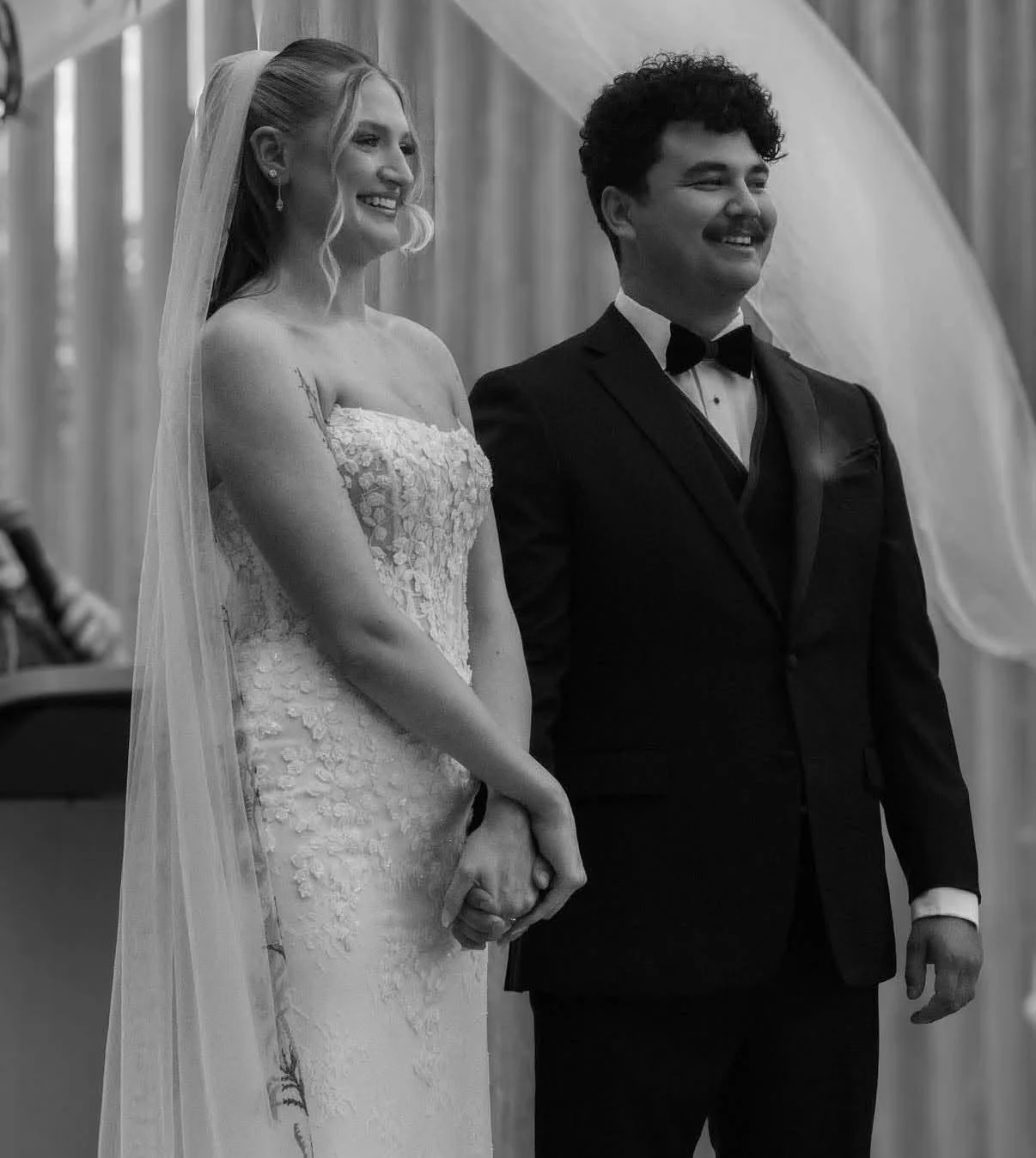 A bride and groom holding hands at their wedding ceremony, smiling and standing in front of a curtain. The bride is in a strapless wedding dress with lace details and a veil, and the groom is dressed in a black tuxedo with a bow tie.