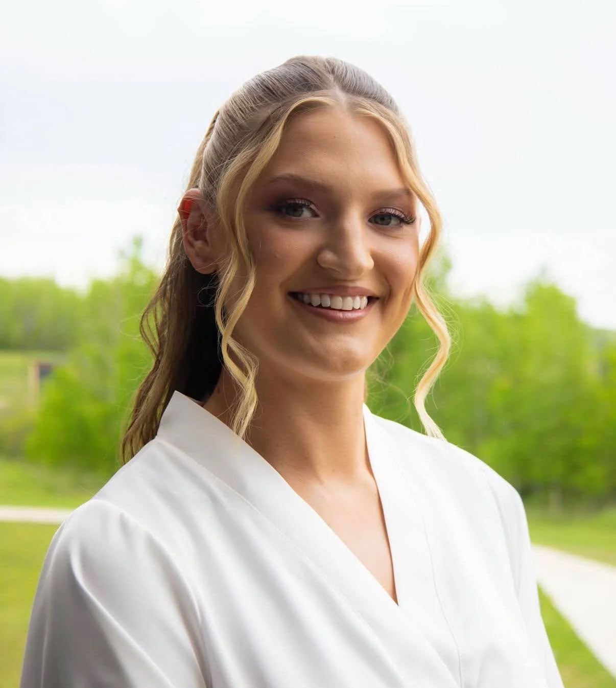 A young woman with blonde hair styled in soft curls, smiling and wearing a white blouse, standing outdoors with a background of trees and a cloudy sky.