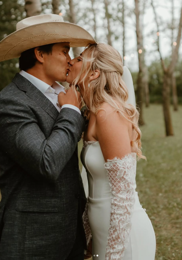 A couple sharing a kiss outdoors at sunset, dressed in wedding attire, with trees in the background.