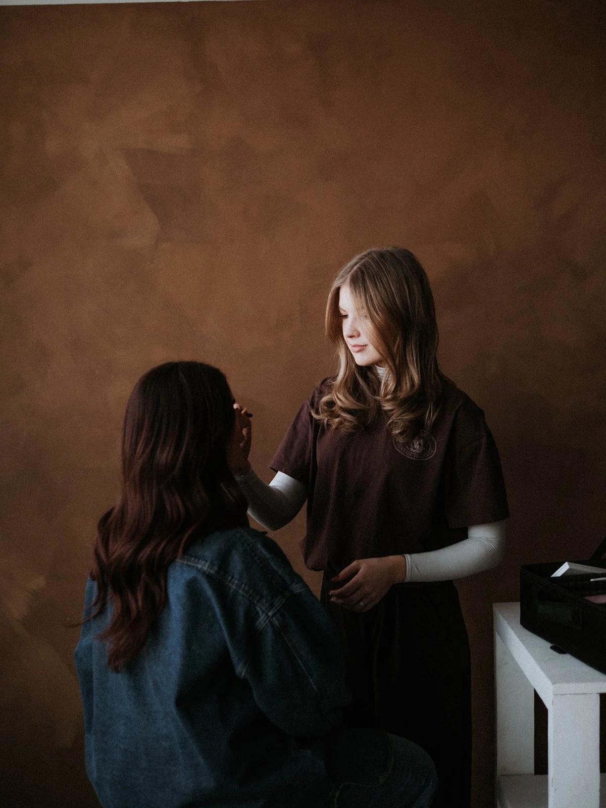 A woman with wavy blonde hair wearing a dark brown shirt applies makeup to a seated woman with long brown hair and glasses at a makeup station.