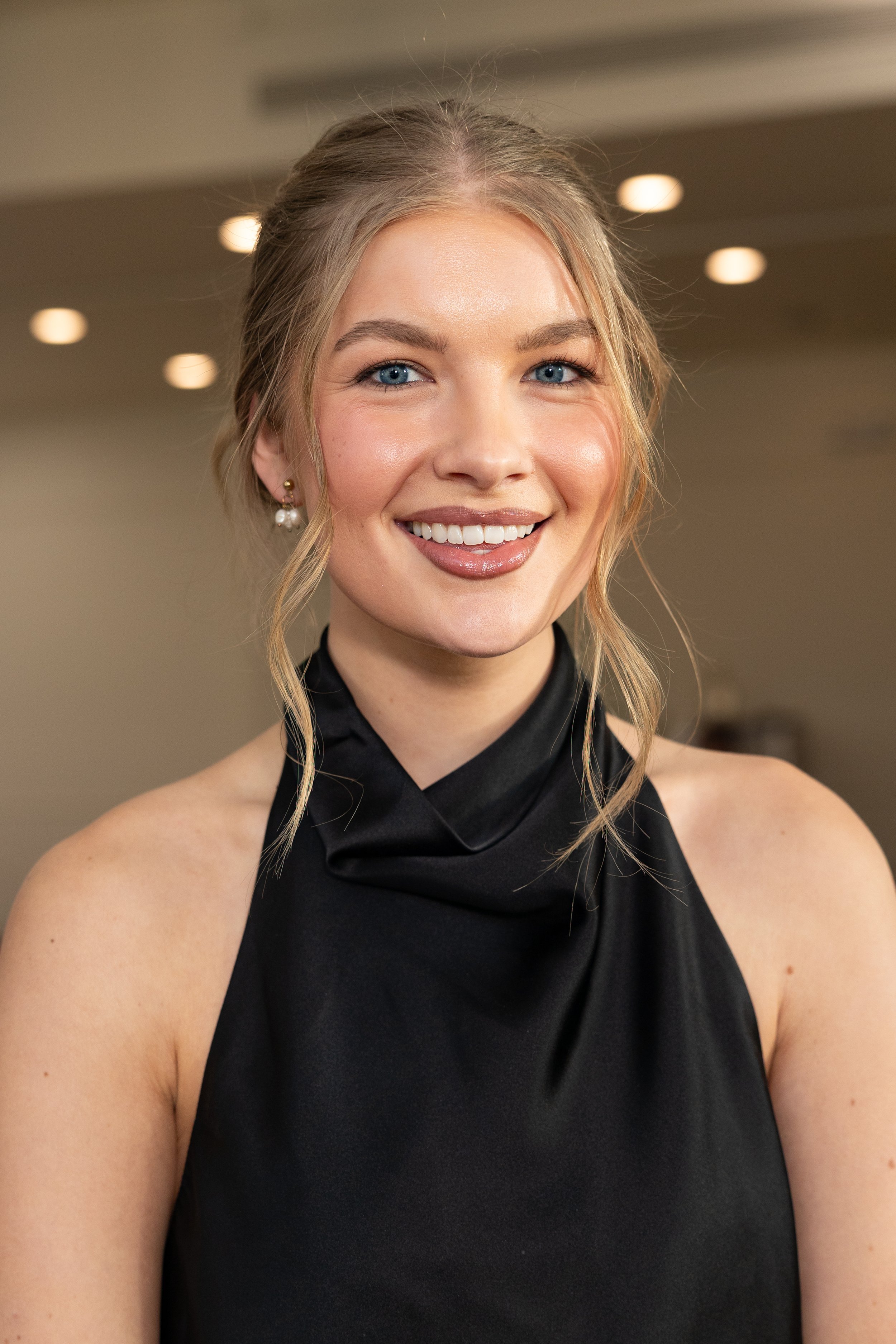 Close-up photo of a young woman with blonde hair styled in a loose updo, wearing a black high-neck satin dress or top, and pearl earrings, smiling with blue eyes in an indoor setting with blurred ceiling lights.