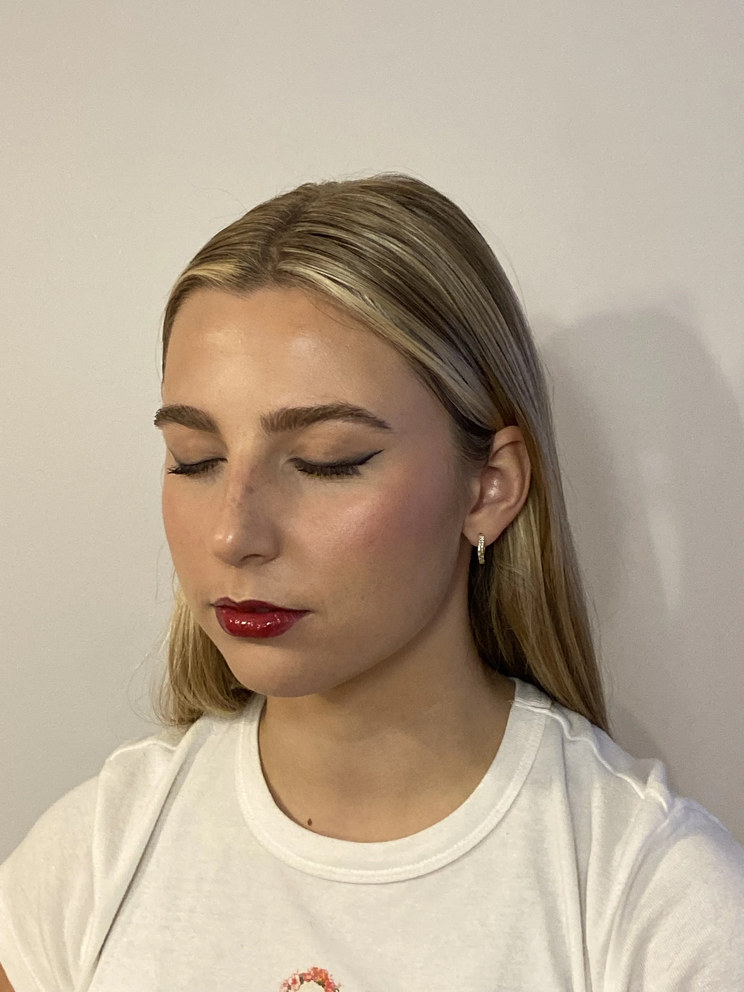 A woman with blonde hair and closed eyes, wearing makeup and a white shirt, standing against a plain wall.