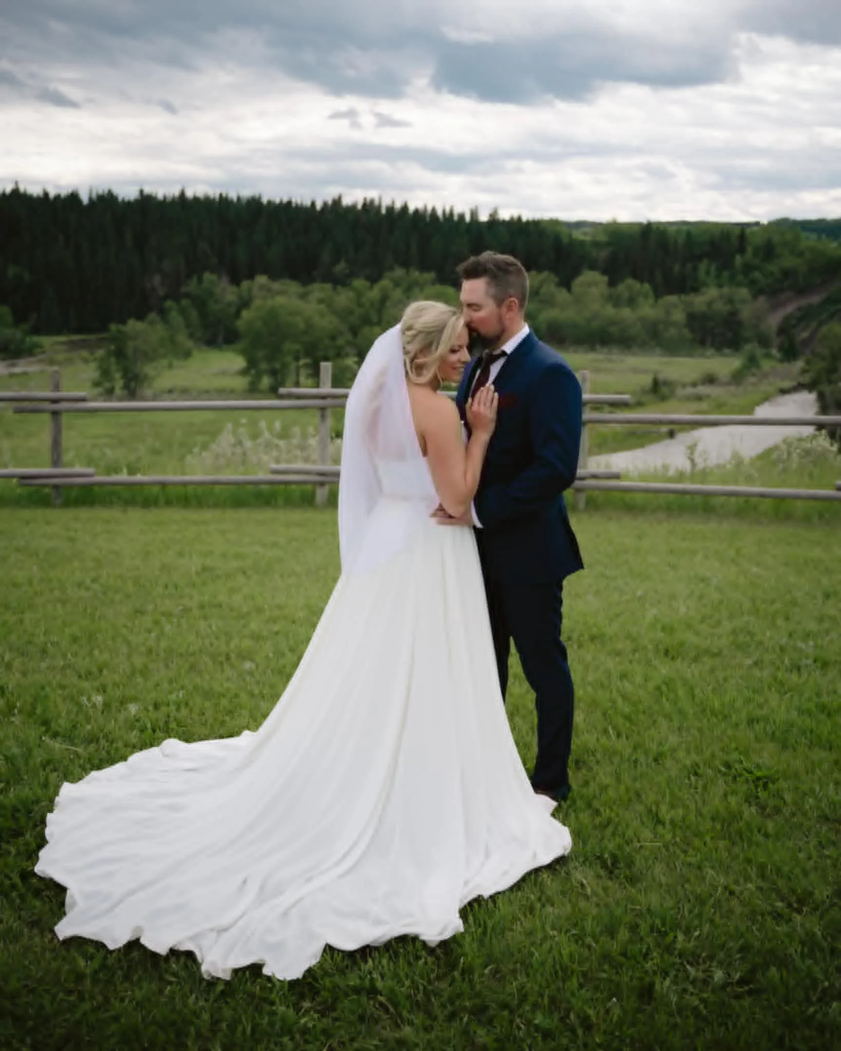A bride and groom embracing outdoors on a grassy field with a wooden fence and green trees in the background. The bride is wearing a white wedding dress and veil, and the groom is in a dark suit. Overcast sky overhead.