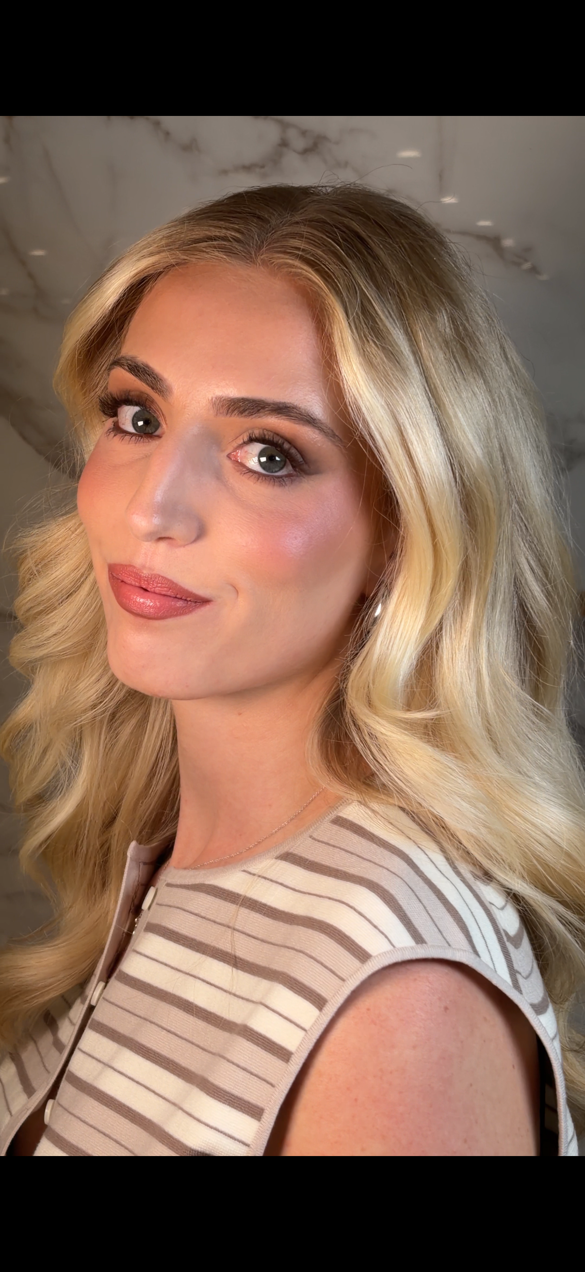 Close-up of a woman with blonde wavy hair, light makeup, wearing a beige and white striped sleeveless top, standing in front of a blurred marble background.