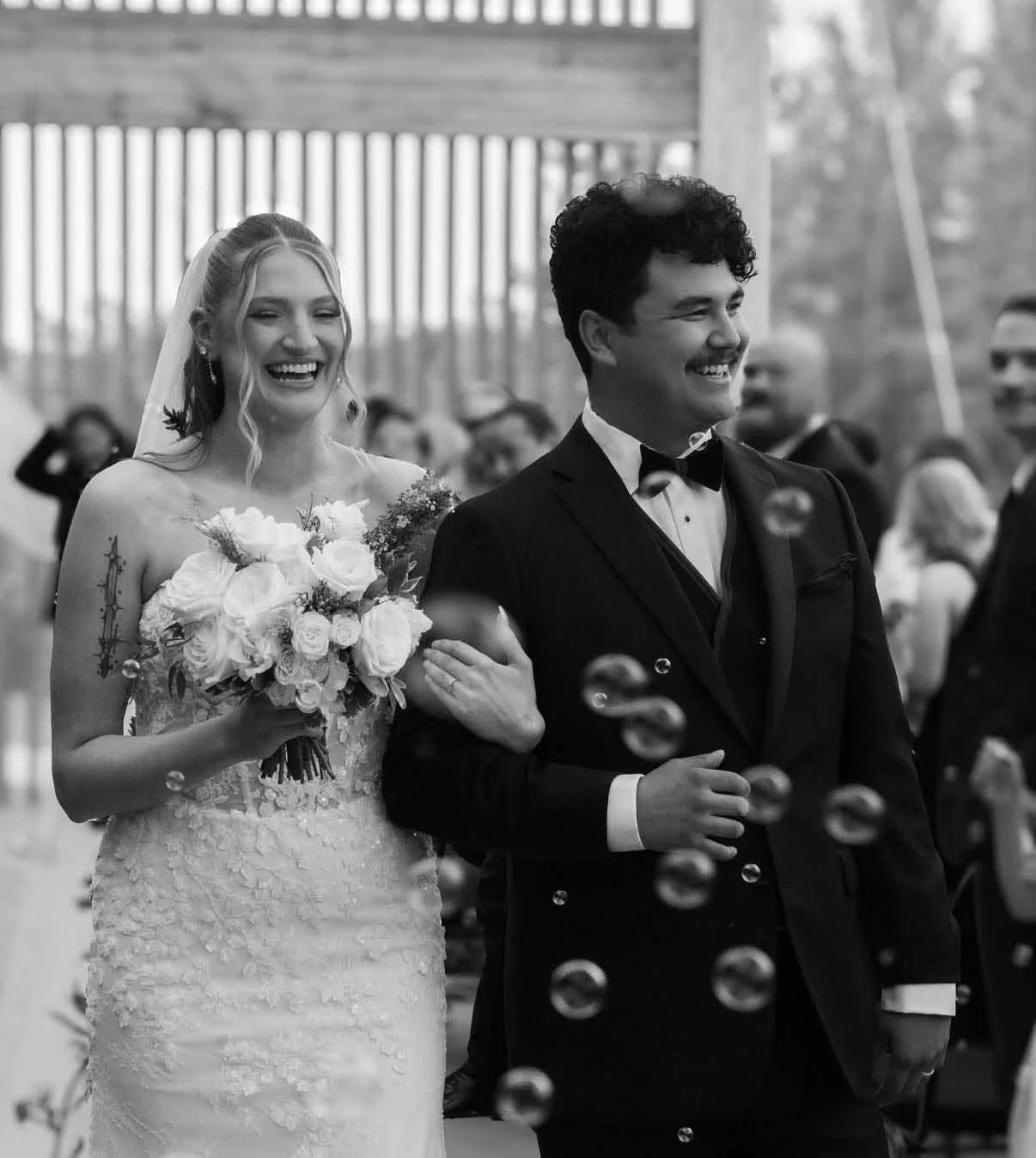 Black and white photo of a bride and groom at their wedding. The bride is smiling and holding a bouquet of flowers, while the groom is dressed in a tuxedo and smiling.