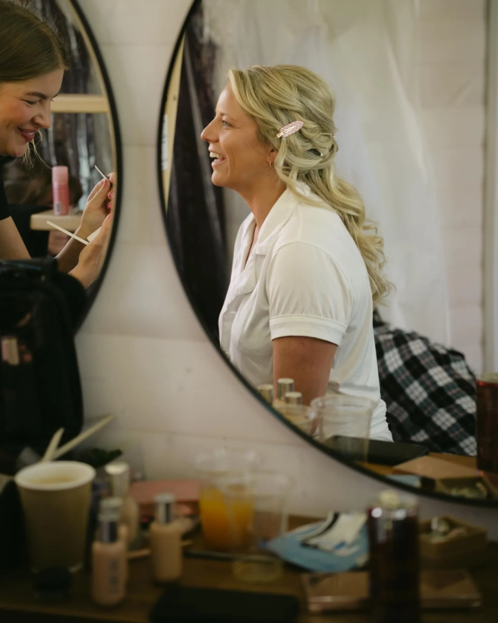 A woman with blonde hair styled with pink hair clips is smiling as she gets her makeup done by a makeup artist. The scene is viewed through a mirror reflection in a room with a cluttered table holding various makeup and beauty products.