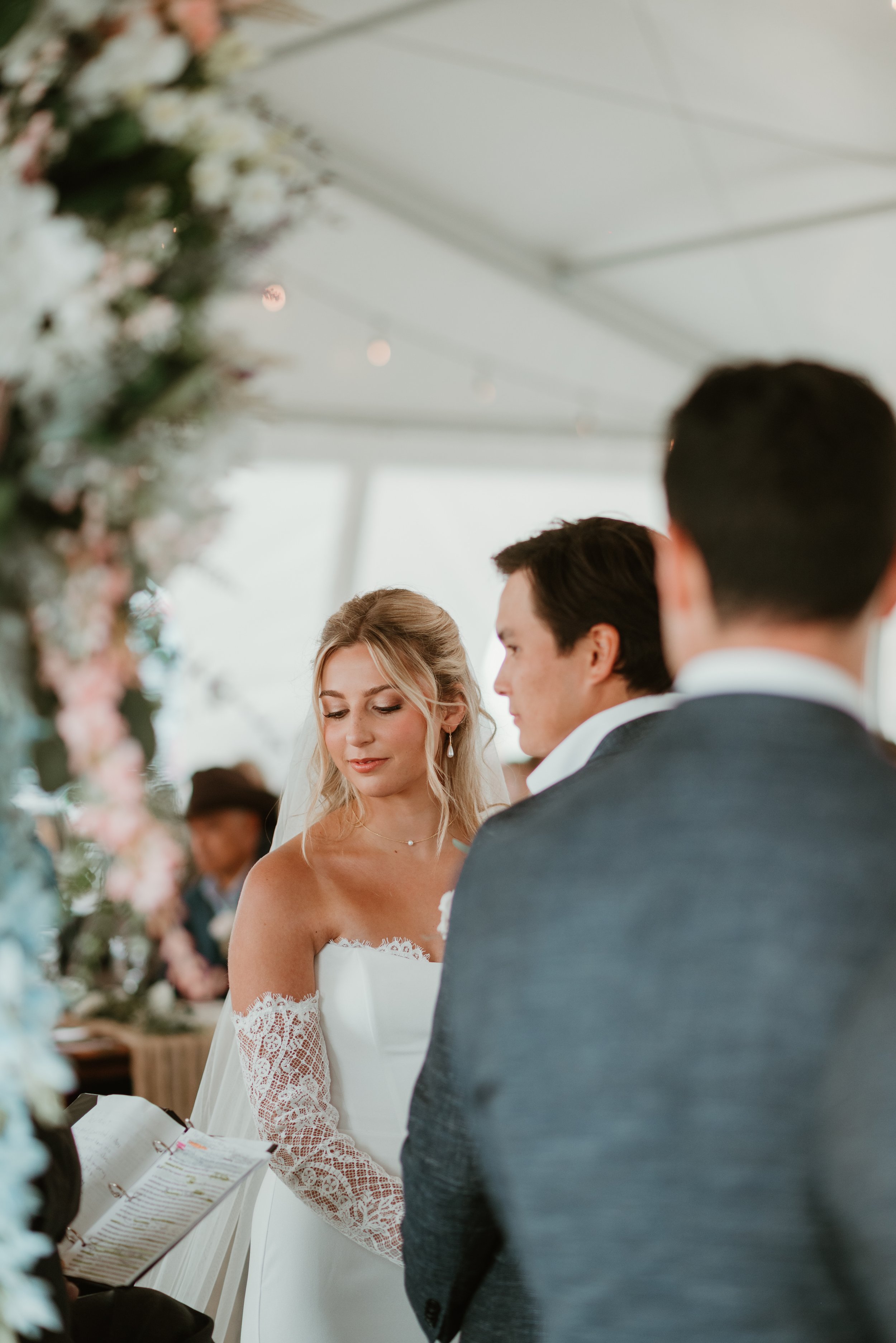 A bride and groom standing at the altar during their wedding ceremony, with a floral arrangement in the foreground and guests in the background.