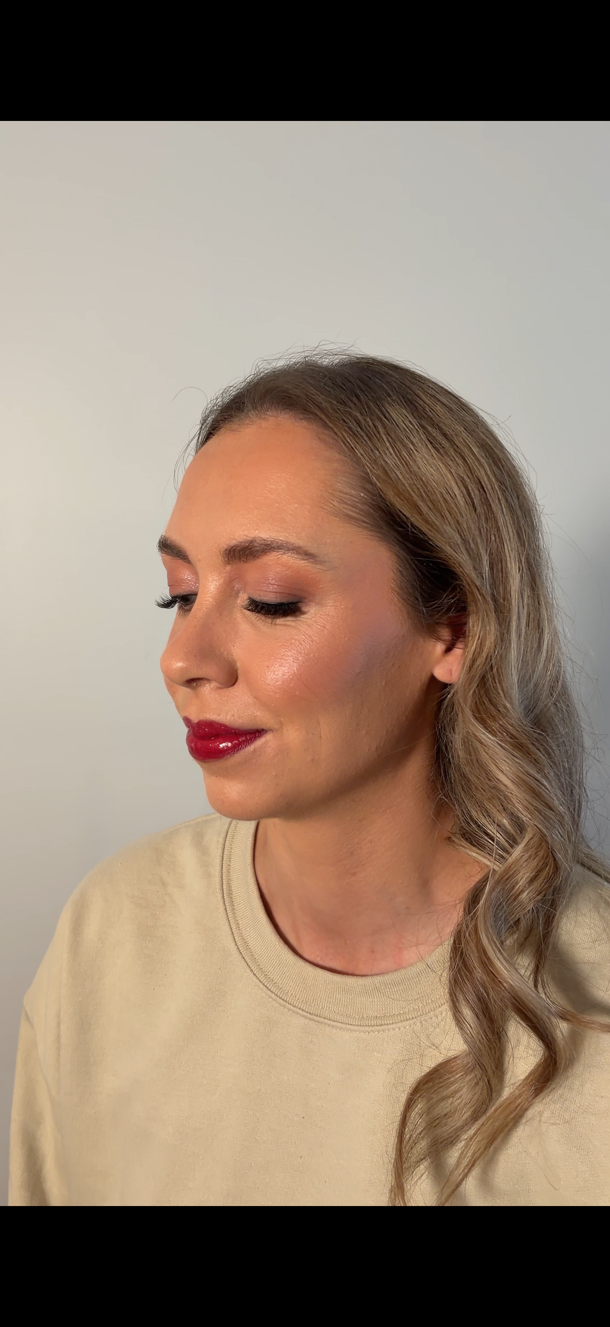 A woman with wavy, blonde hair styled to one side, wearing makeup including red lipstick and eye makeup, dressed in a beige shirt, standing against a plain white wall.
