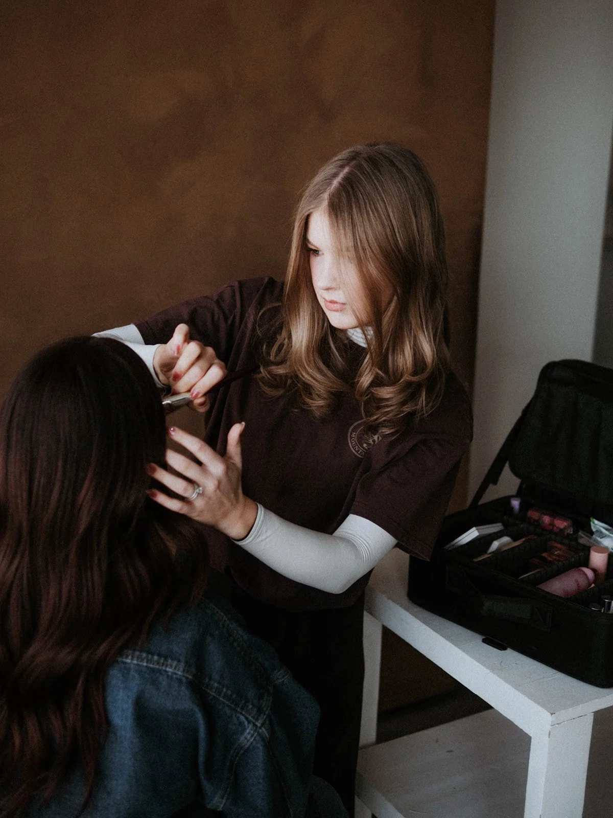 A woman with long, wavy blonde hair is applying makeup to another woman with dark hair, seated in front of her. The makeup artist is using a brush, and there is a makeup case with various cosmetics on a white table beside them.