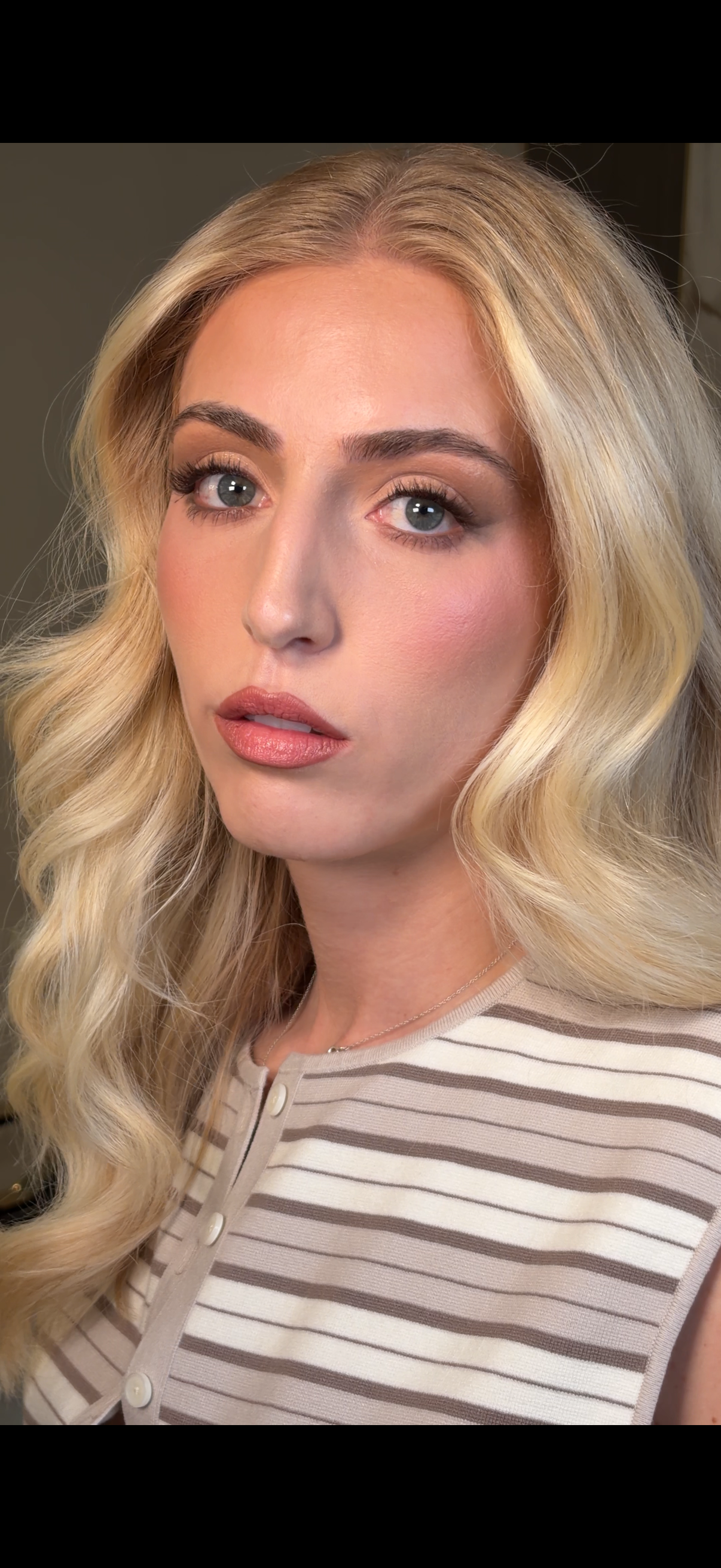 Close-up of a woman with long blonde wavy hair, blue eyes, and natural makeup wearing a beige and white striped top.