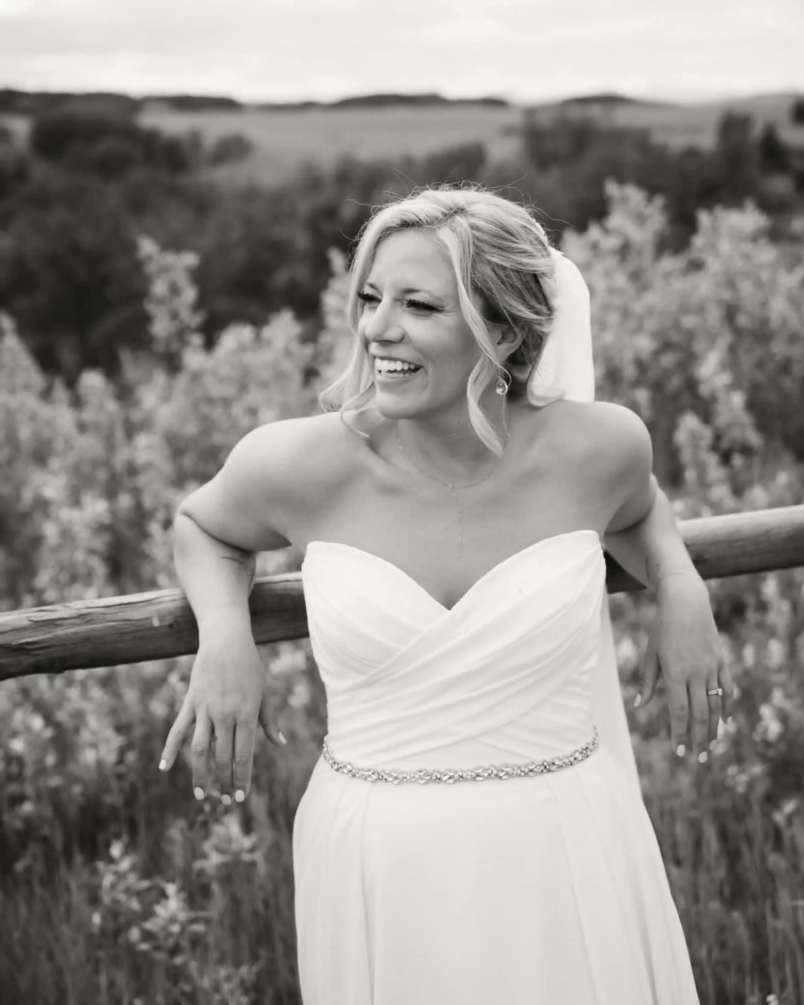 A woman in a strapless wedding dress, smiling and looking to the side, outdoors with a blurred natural background.