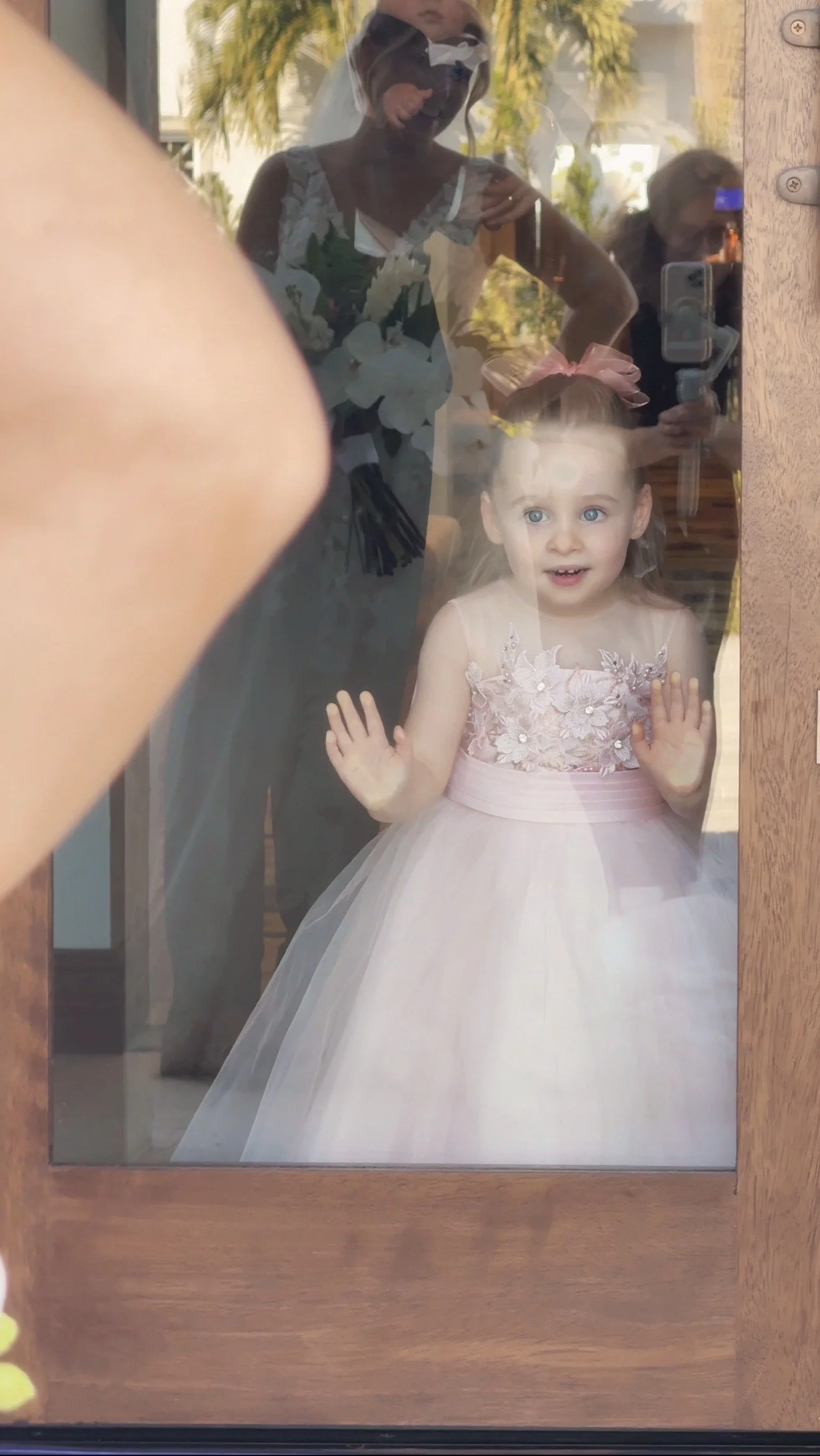 A young flower girl in a pink dress with floral embroidery and a pink bow in her hair, standing behind a glass door, pressing her hands against it and looking outside with a curious expression.
