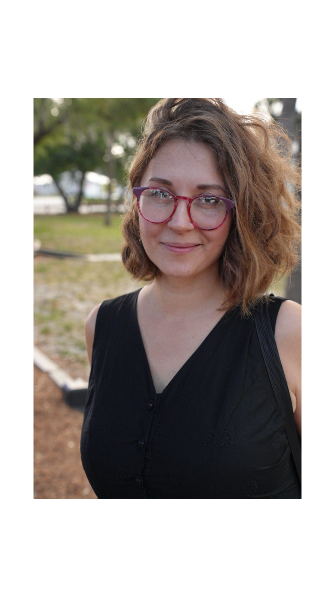 A young woman with curly, light brown hair and glasses standing outdoors in a park with trees and grass in the background.