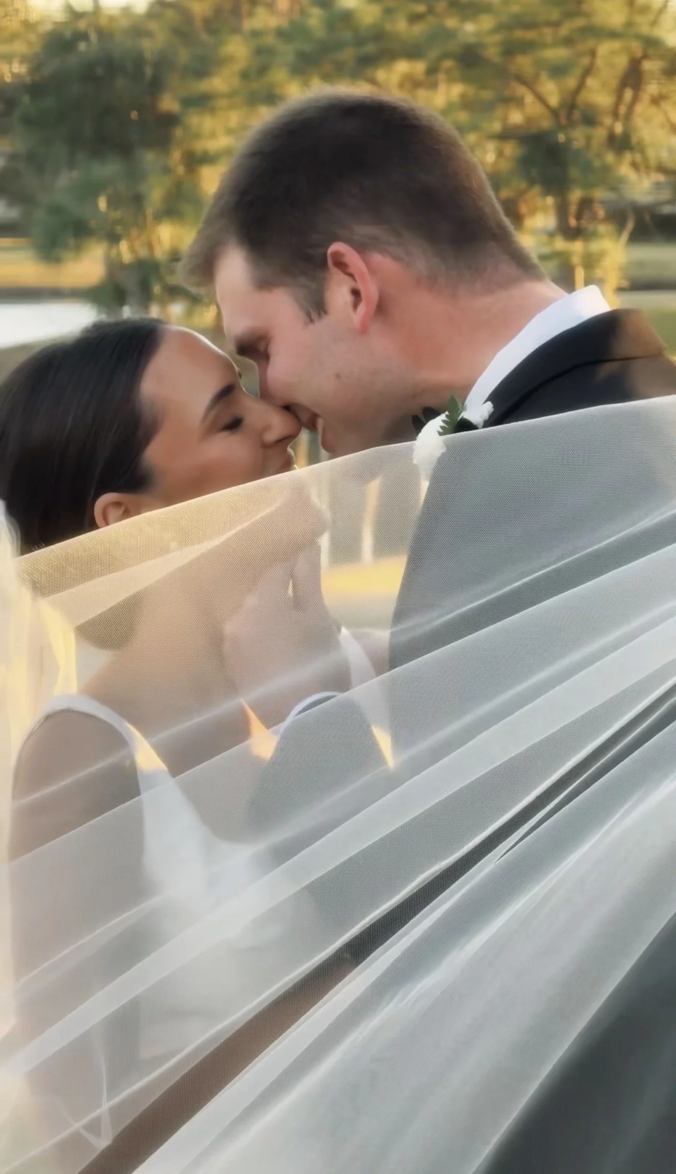 A bride and groom sharing a close moment, forehead to forehead, outdoors with trees and a pond in the background, sunlight casting a warm glow.