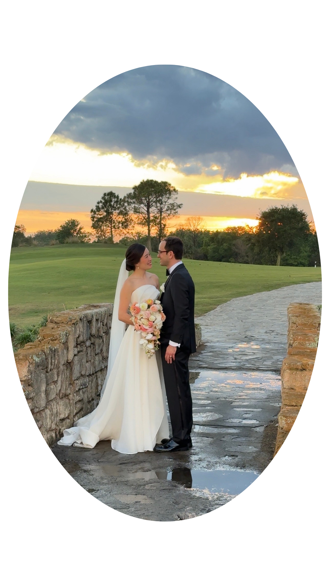 A bride and groom standing on a stone pathway on their wedding day at sunset, with the bride holding a large bouquet of pink and white flowers, both smiling and looking at each other.