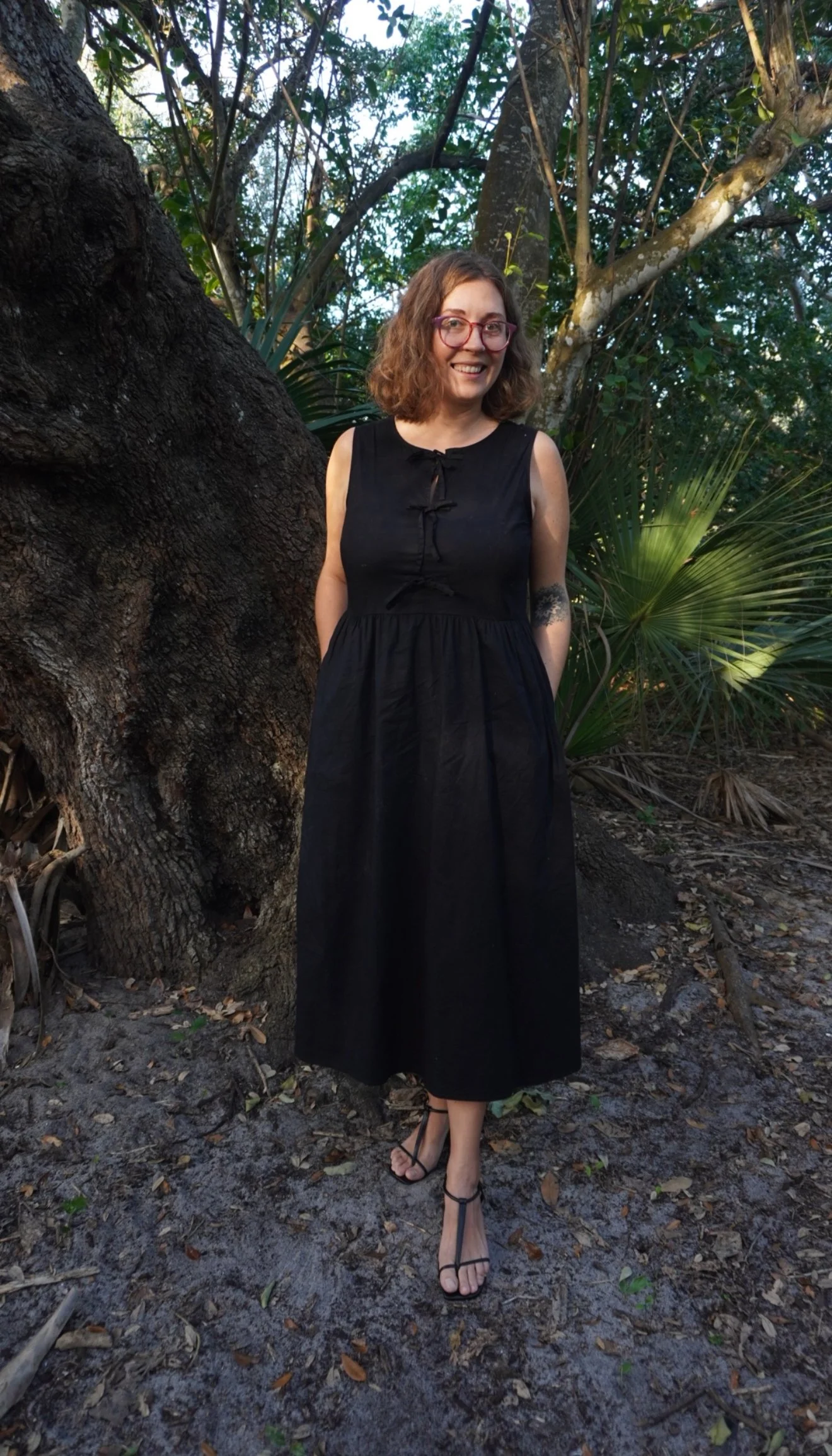 A woman with shoulder-length curly brown hair, glasses, and a tattoo on her left arm stands outdoors in front of a large tree and lush greenery, wearing a sleeveless black dress and black sandals. She is smiling and looking at the camera.