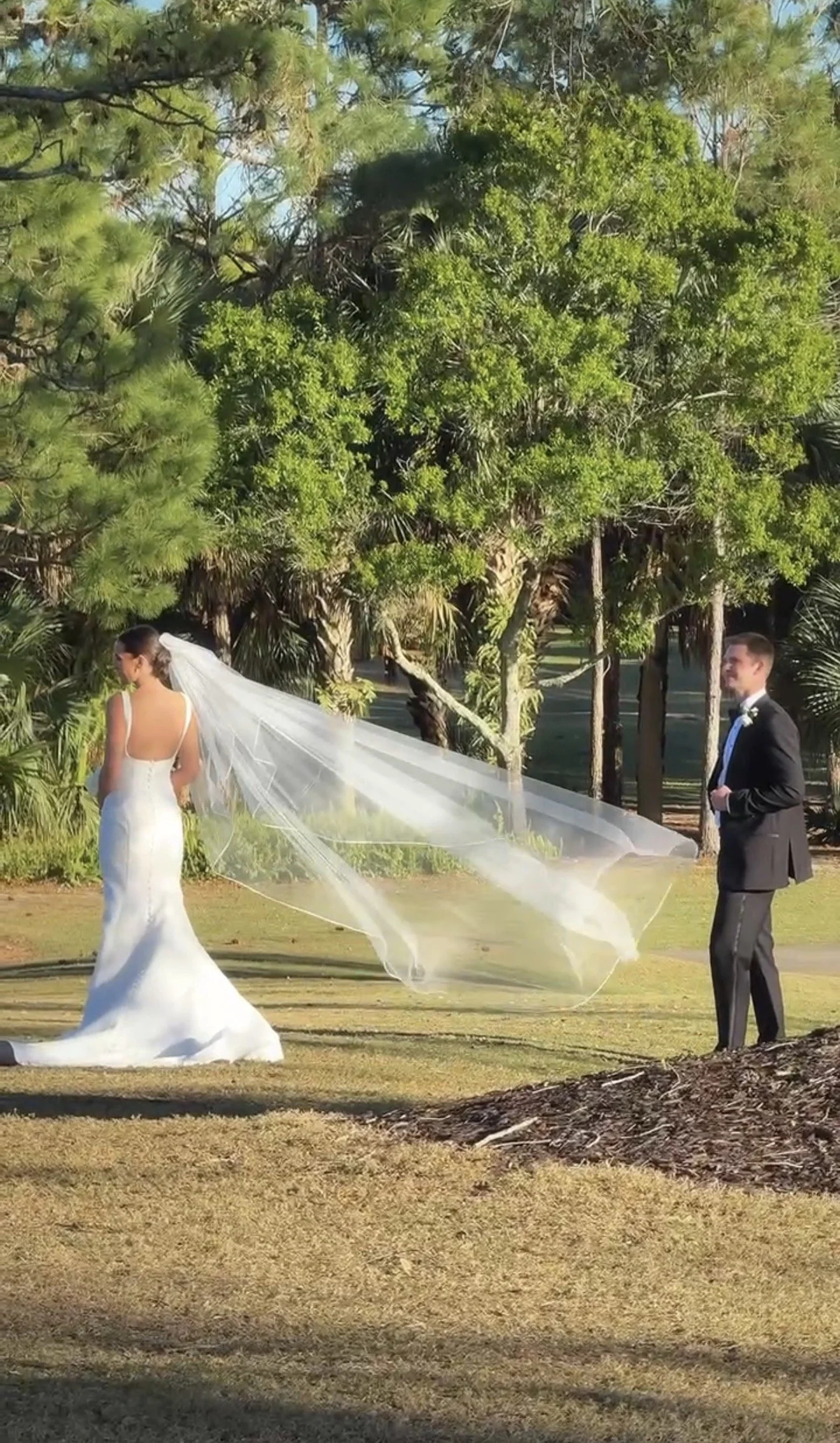 A bride and groom outdoors during a wedding ceremony. The bride is in a white gown with a long veil, and the groom is in a black tuxedo. Tall green trees provide a natural backdrop.