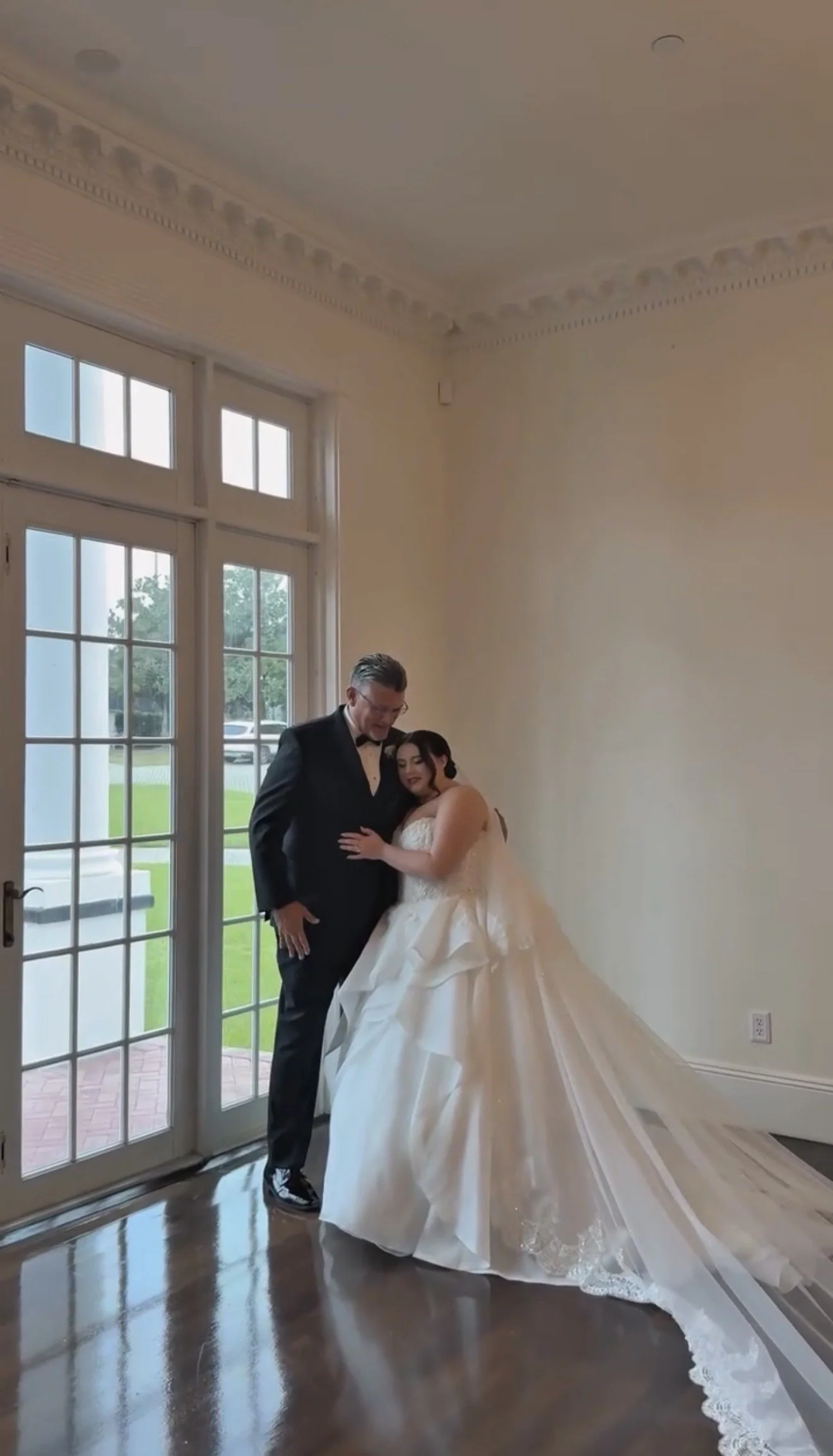 A bride and her father stand together by a large glass door in a spacious room with ornate ceiling moldings. The bride is wearing a white wedding gown, and the groom is wearing a black suit and bow tie.