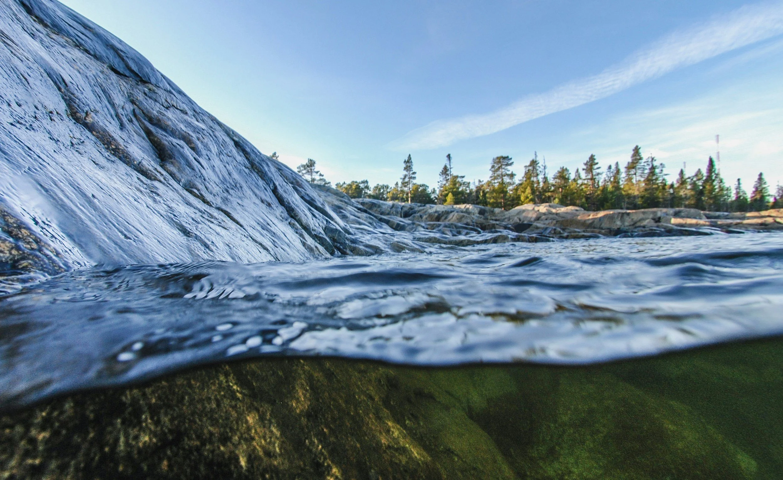 Blick unter Wasser an einem Fluss mit glatter, schräg ansteigender Felswand und Bäumen im Hintergrund bei klarem Himmel.