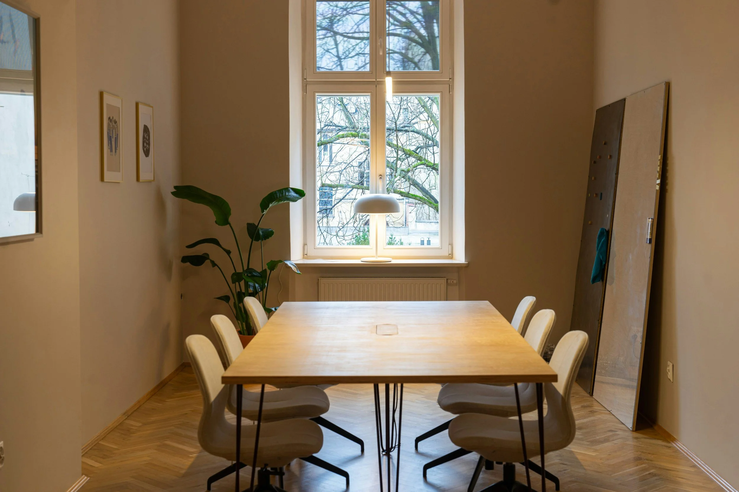 Ein Esszimmer mit einem rechteckigen Holztisch und sechs weißen Stühlen. Im Hintergrund ein großes Fenster mit Blick auf Baumzweige. An der Wand sind zwei Bilder in goldenen Rahmen und eine große Tafel oder ein Brett. Es gibt eine weiße Lampe über dem Fenster und eine grüne Zimmerpflanze neben dem Tisch.