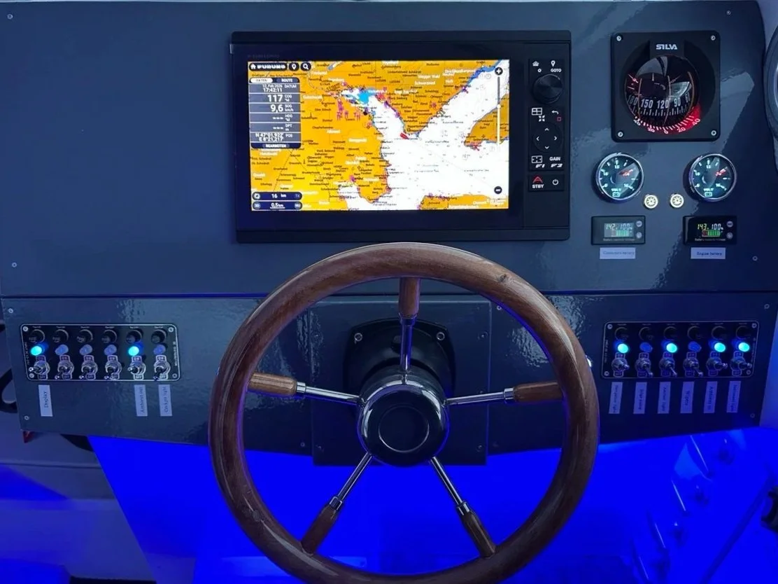 Navigation and control panel for a boat or yacht, featuring a GPS display showing a map, gauges, switches, and a wooden steering wheel.