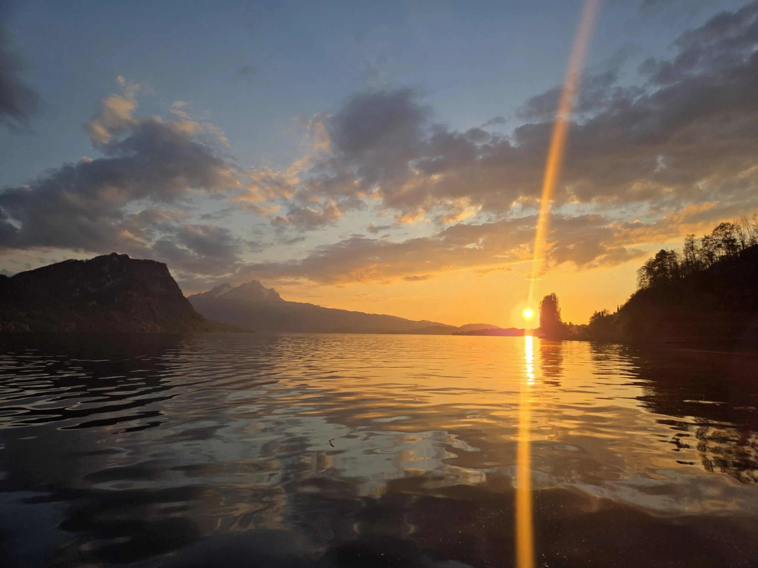 Sunset over a lake with mountains in the background, a rainbow in the sky, and trees silhouetted in the foreground.