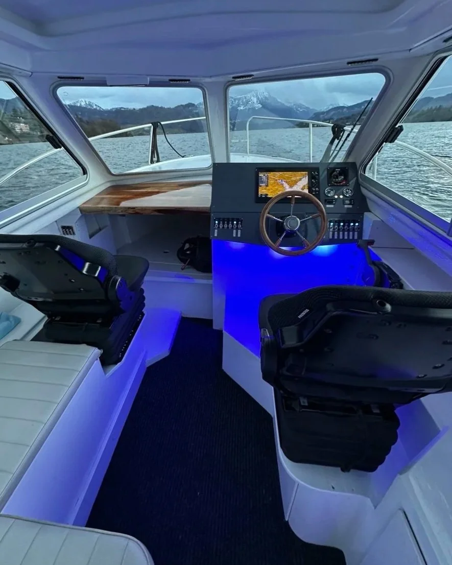 View of a boat's cockpit with two captain chairs, a central steering wheel, and navigation display, with water and mountains in the background.