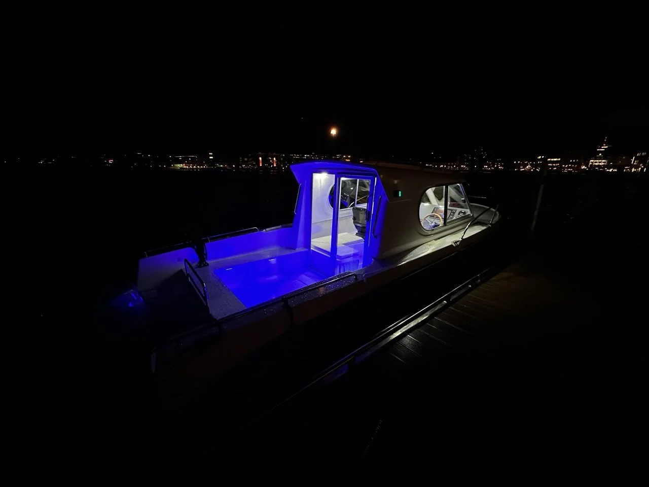 A boat docked at night with blue lighting illuminating the boat's deck and interior, city lights in the background under a dark sky.
